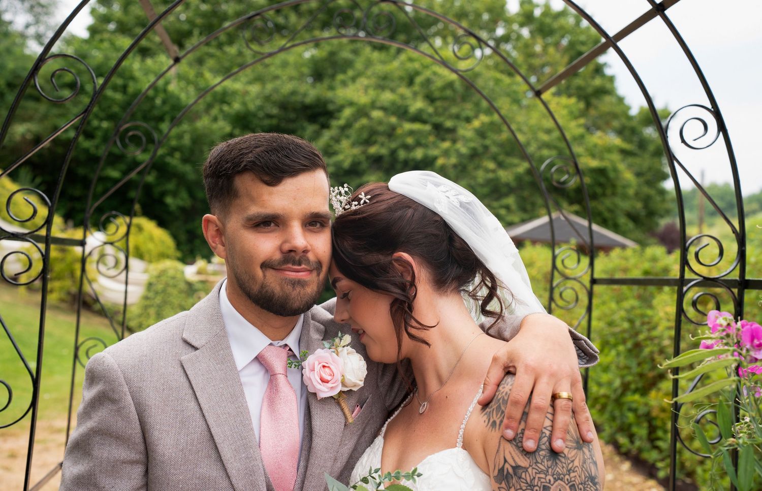 Bride and groom embracing tenderly under decorative iron arch in garden, groom in grey suit with pink tie.