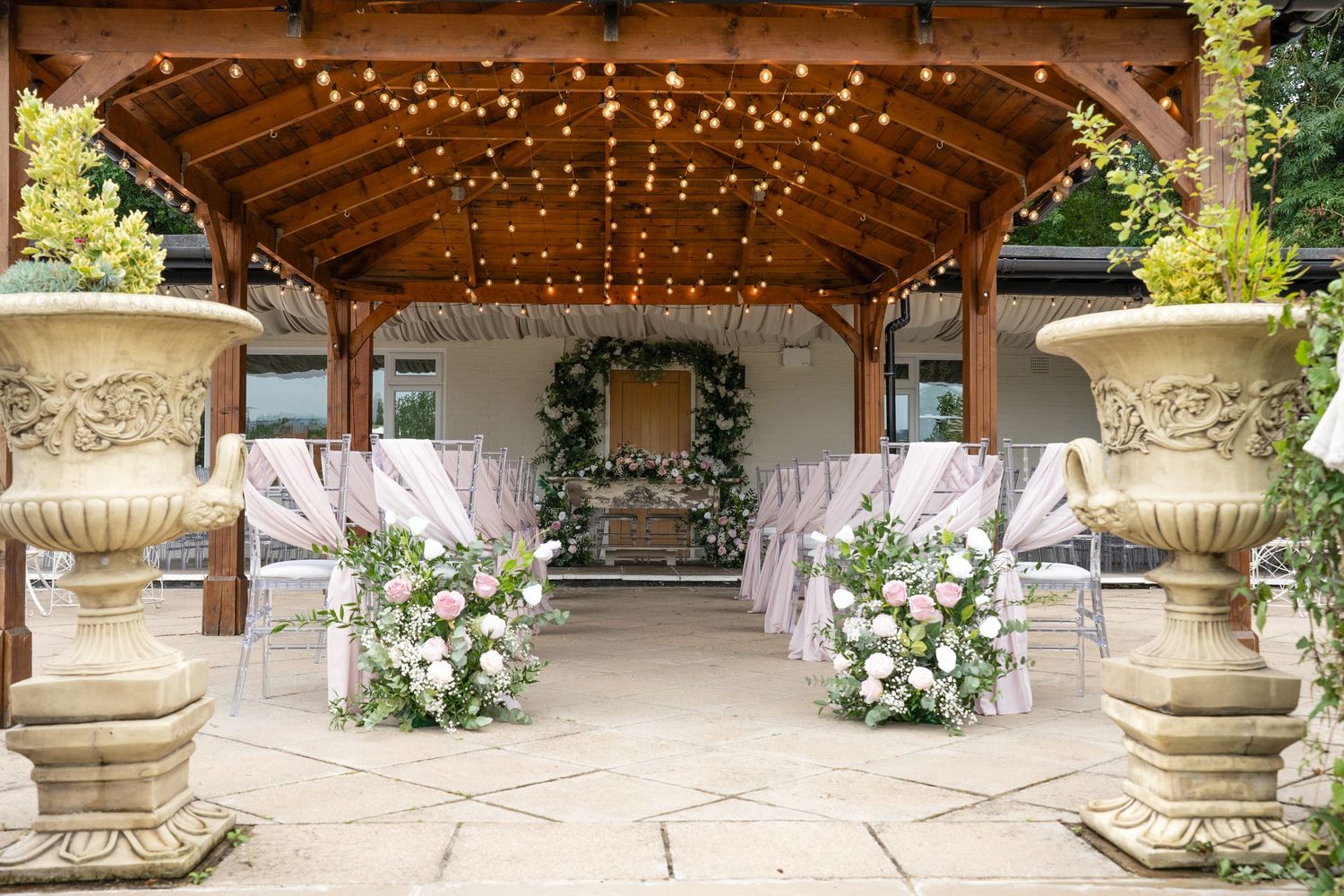Outdoor wedding ceremony venue with fairy lights, floral arrangements and decorated chairs.