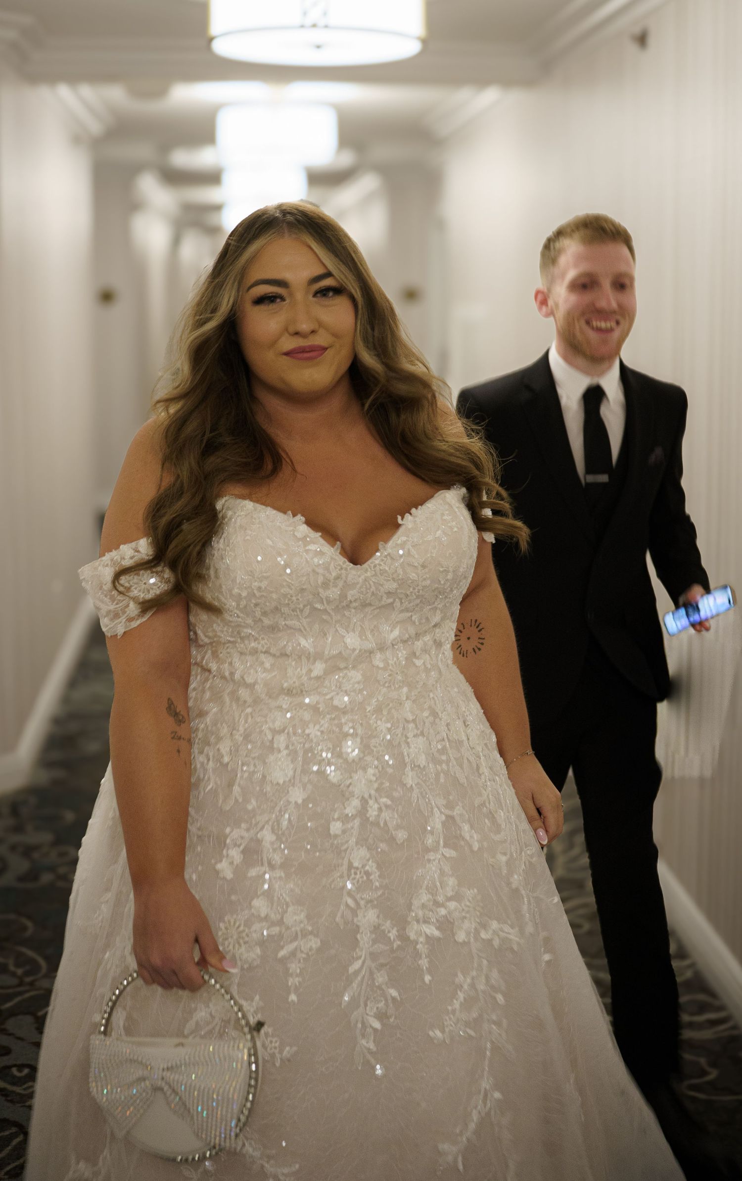 Bride in off-shoulder lace wedding gown walks hotel corridor with smiling groom in black suit.