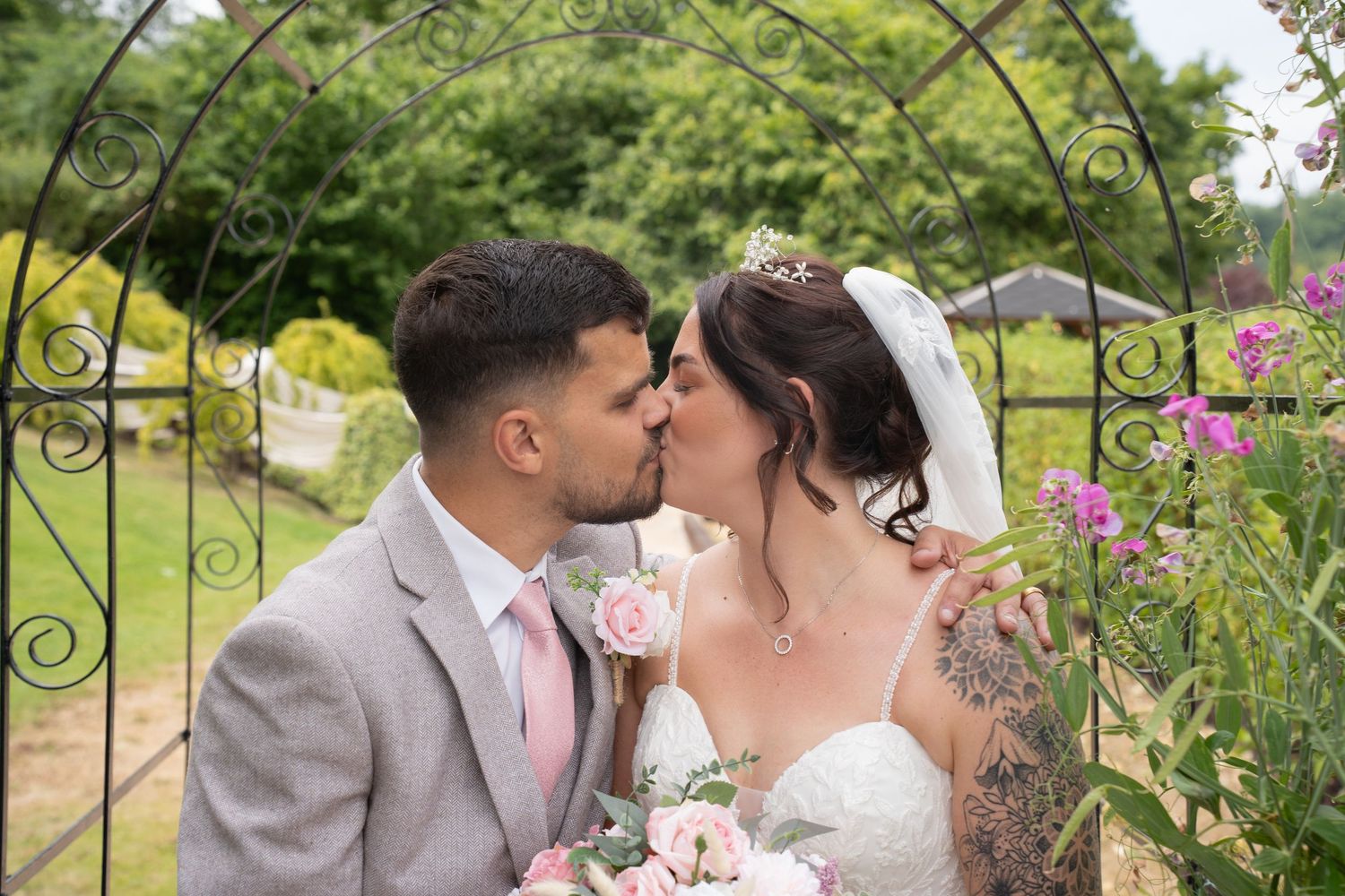 Bride and groom kissing under decorative iron arch in garden wedding setting.