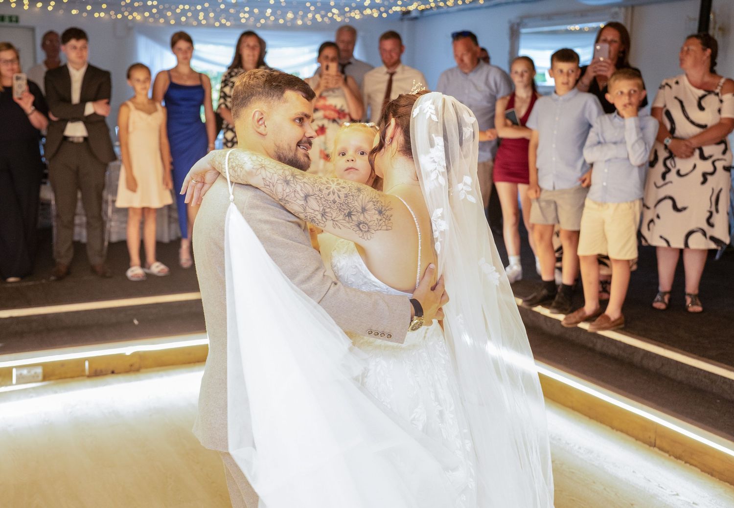 A groom lifts a flower girl during the first dance at a wedding reception with guests watching.
