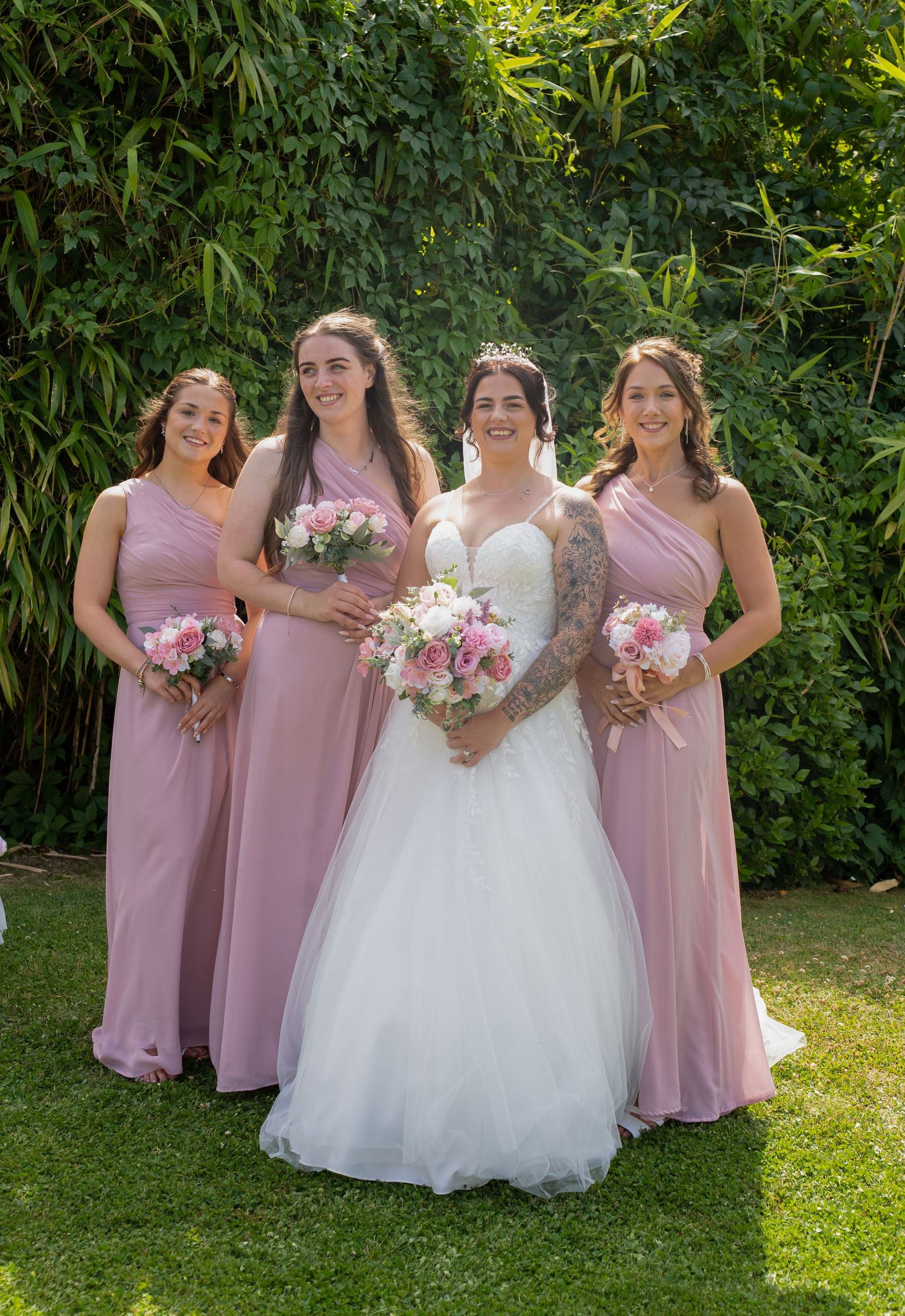 Bride in white gown stands with three bridesmaids wearing matching dusty pink one-shoulder dresses outdoors.