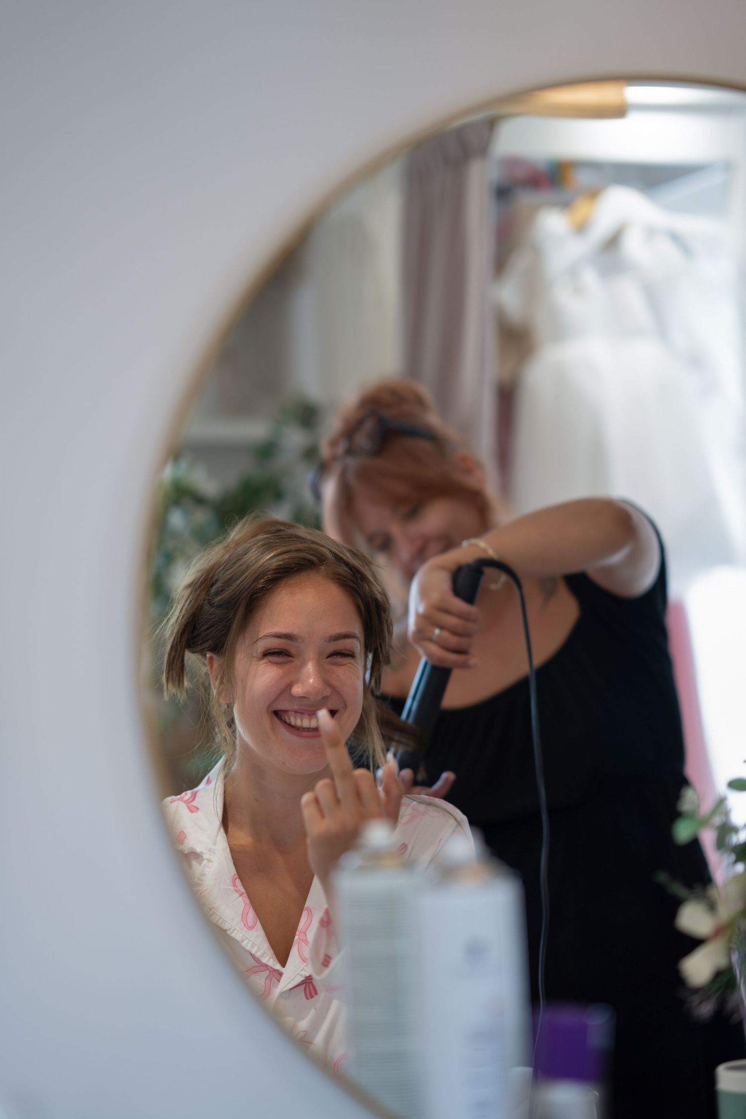 Bride smiling in mirror while hairstylist curls her hair during wedding preparations.