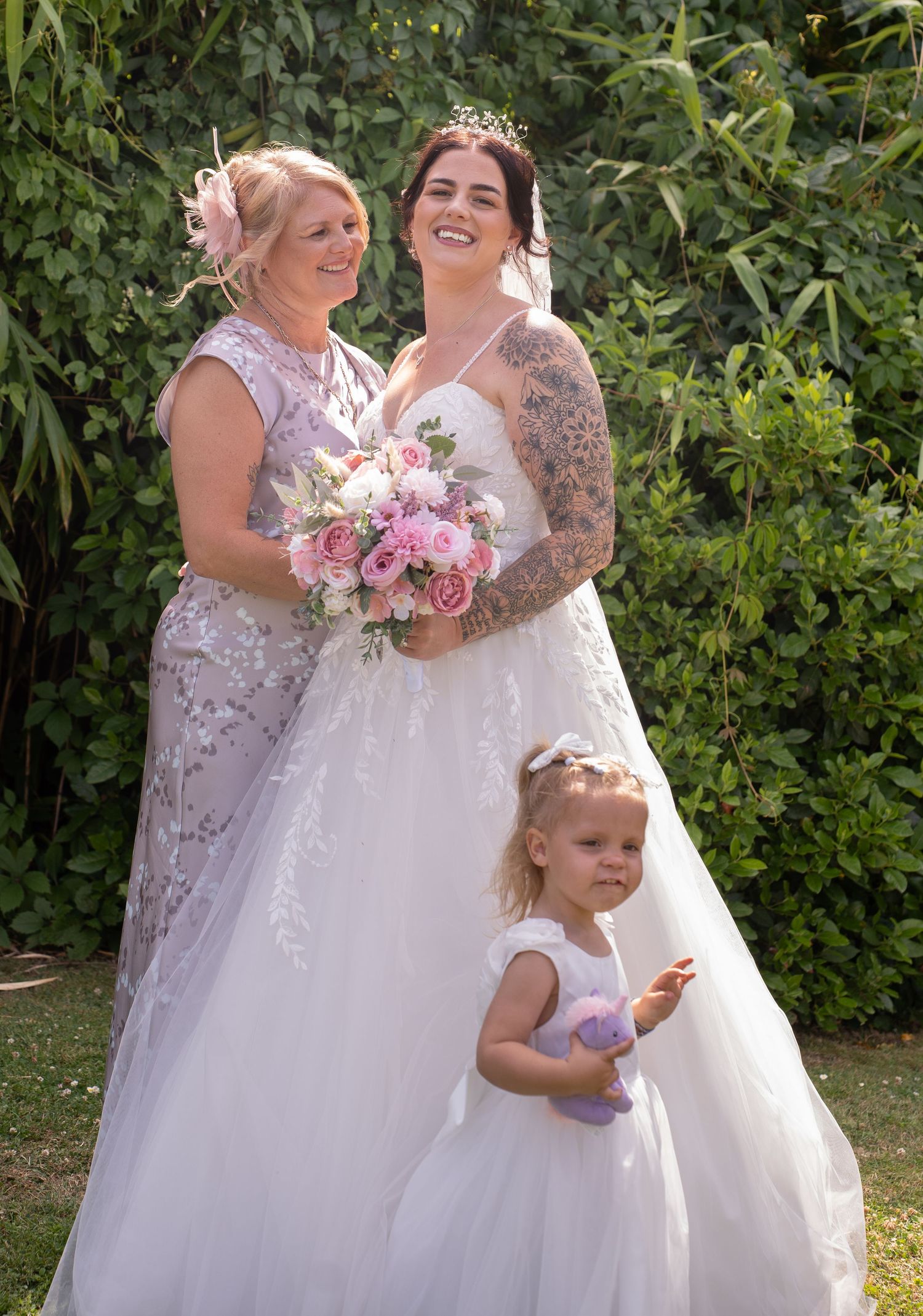 A bride with tattoos holds a pink bouquet posing with an older woman and a flower girl outdoors.