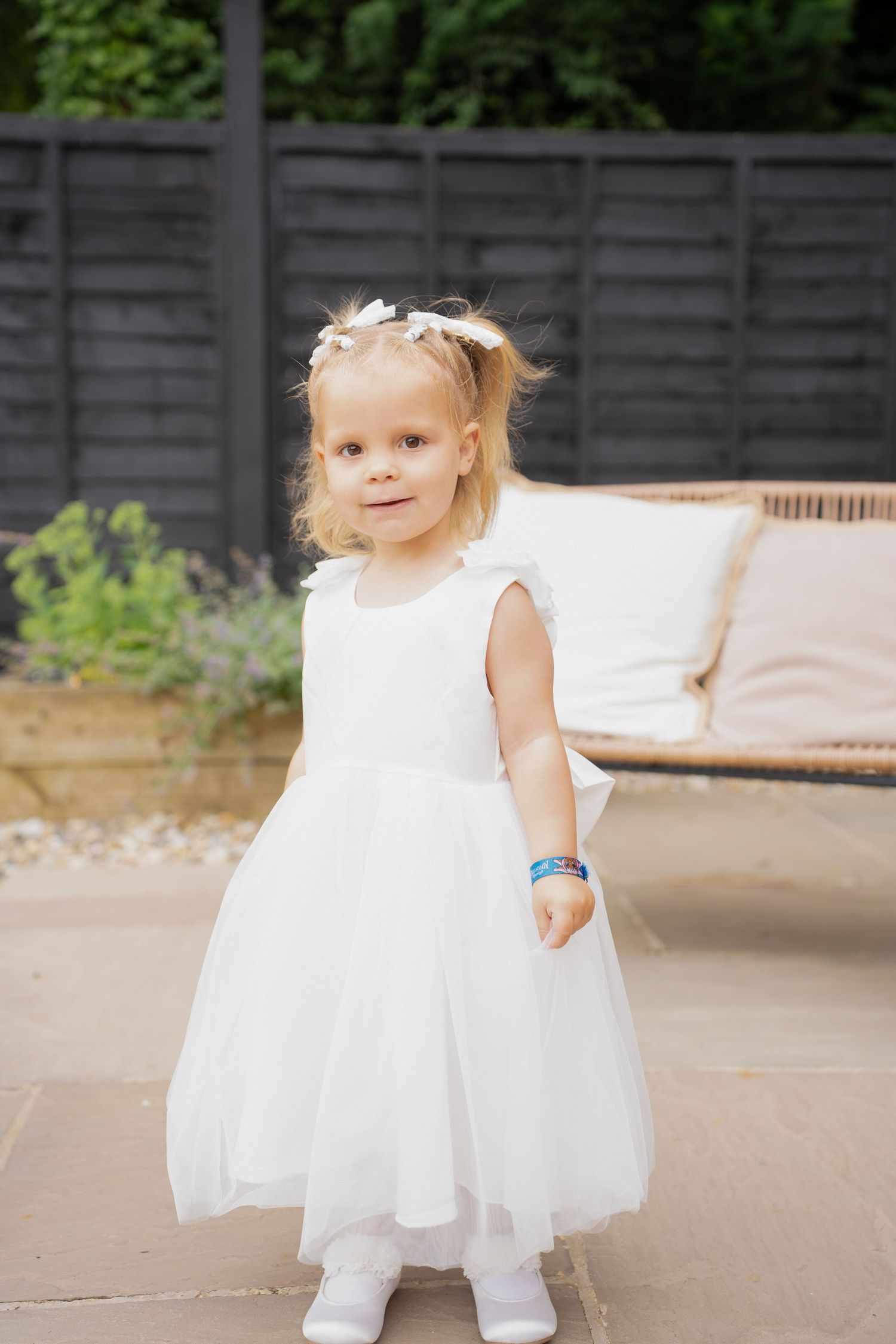 Cute blonde toddler flower girl in white tulle dress standing on patio at outdoor wedding.