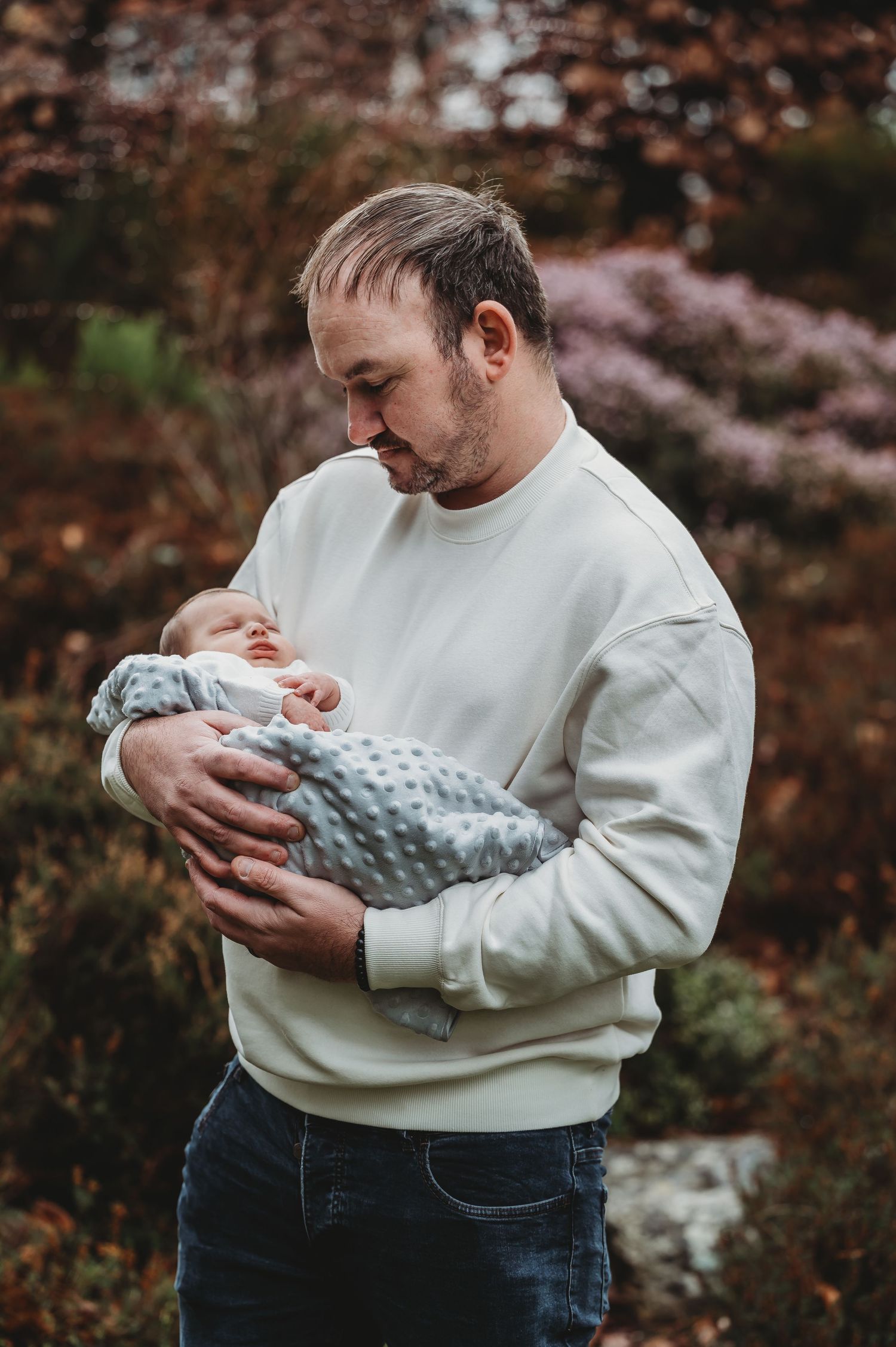 Father tenderly cradling newborn baby outdoors with autumn foliage in background.