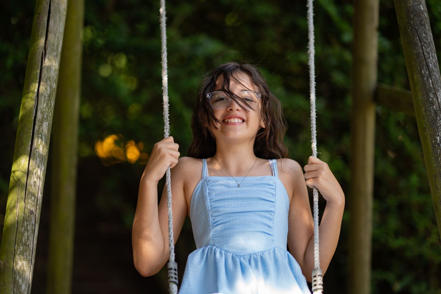 A happy young girl in a light blue dress smiles while swinging on a rope swing outdoors.
