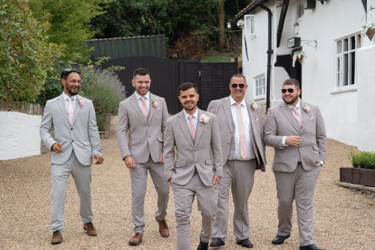 Five groomsmen in matching grey suits with pink ties walking outdoors at wedding venue.