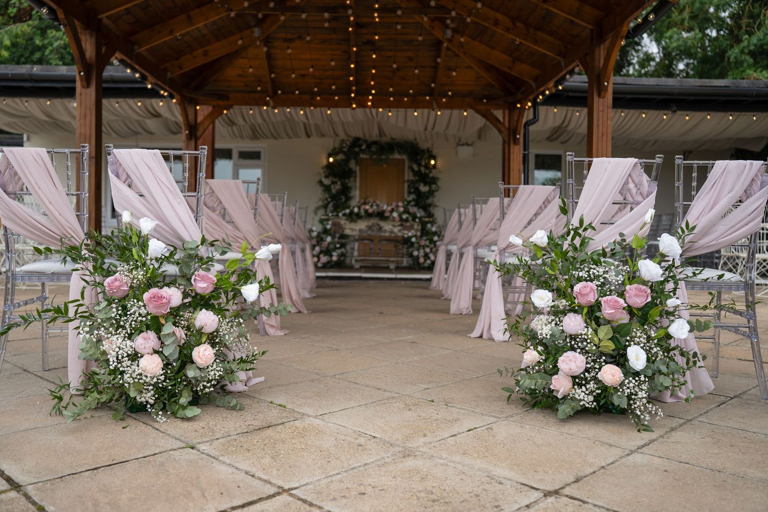 Romantic outdoor wedding aisle with pink floral arrangements and fairy light canopy.