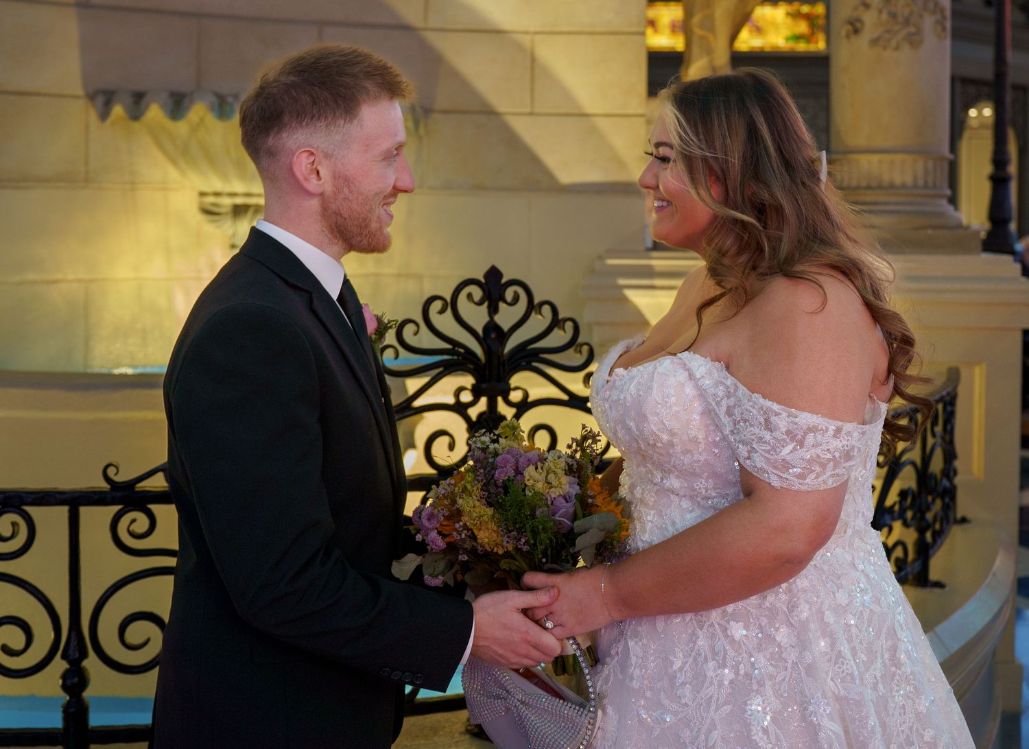 Groom in black suit and bride in lace gown hold hands smiling at Las Vegas wedding venue.