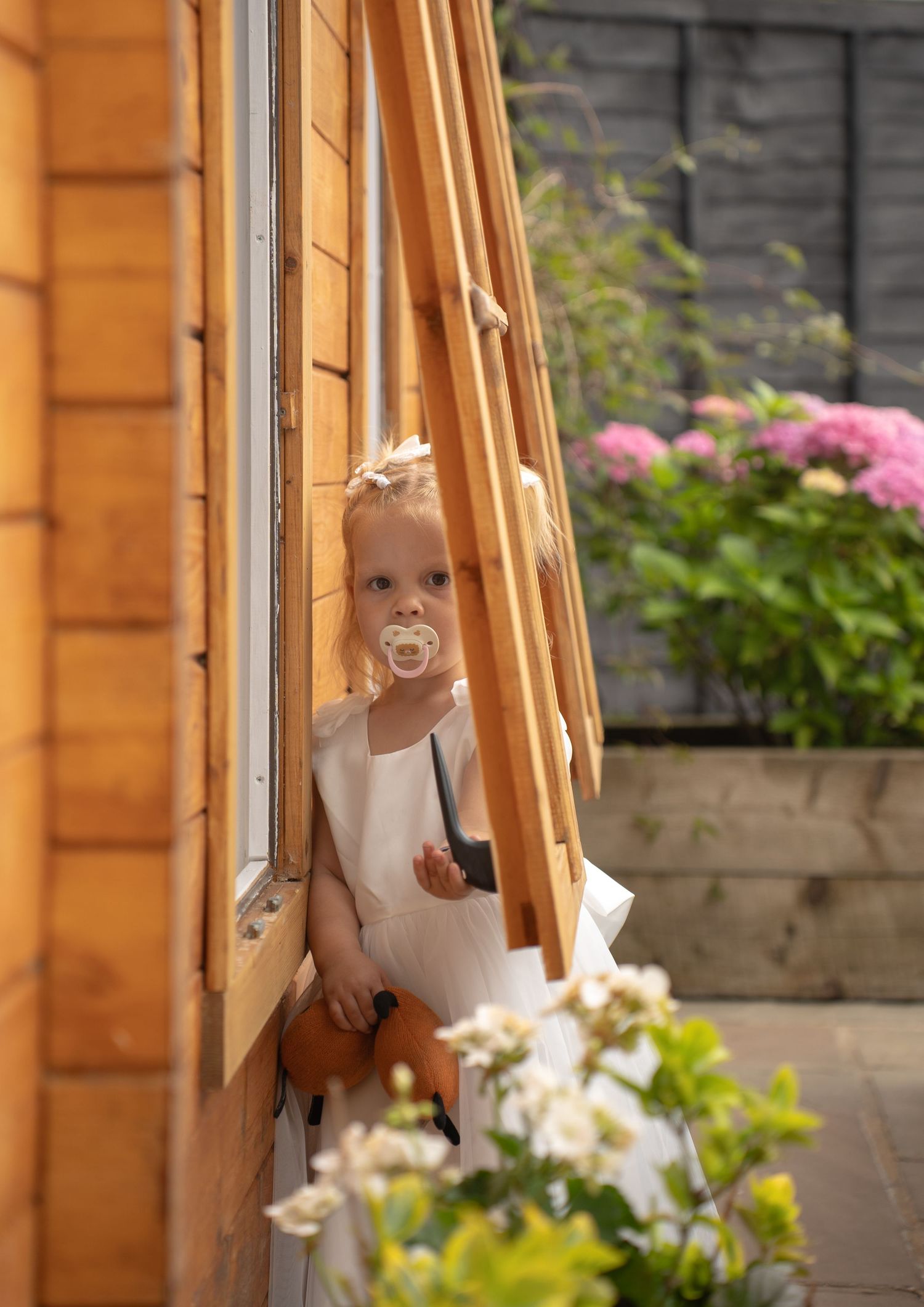 Adorable blonde flower girl in white dress with pacifier peeking around wooden cabin door.