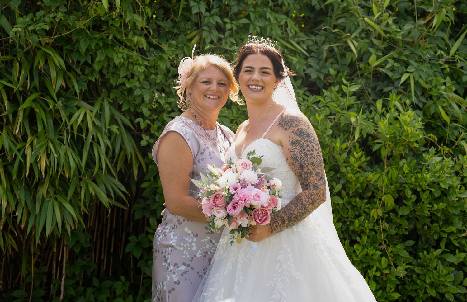 Bride in white gown with tiara holding pink bouquet posing with smiling woman in floral dress outdoors.