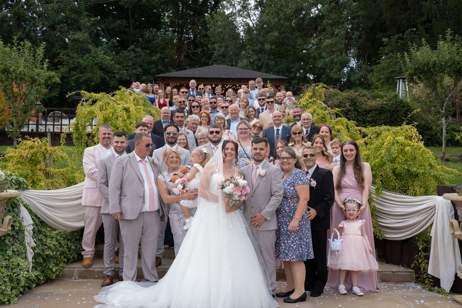 Large wedding group photo with bride and groom surrounded by guests in garden.