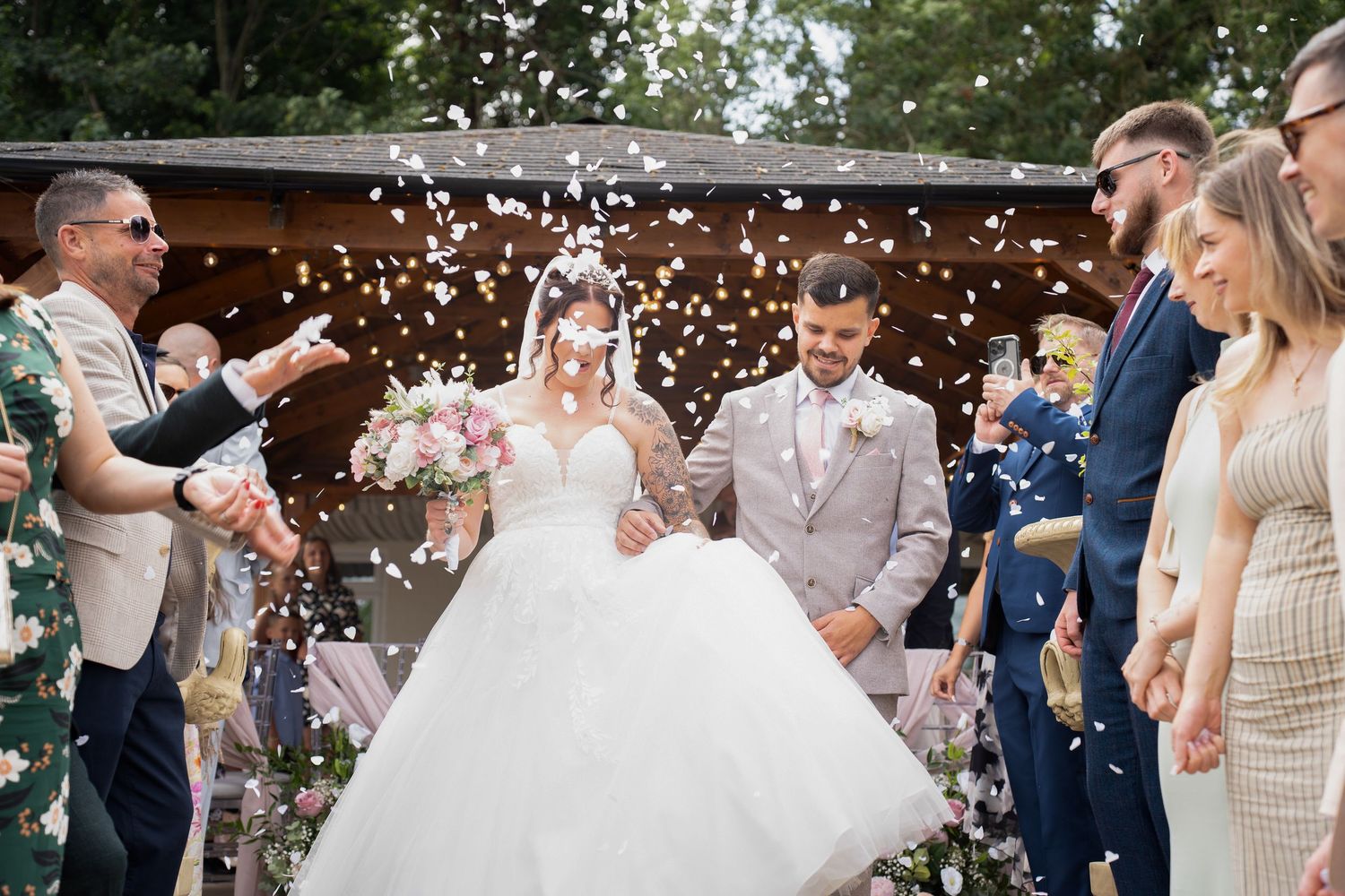 Bride and groom walk through confetti shower surrounded by smiling wedding guests outdoors.