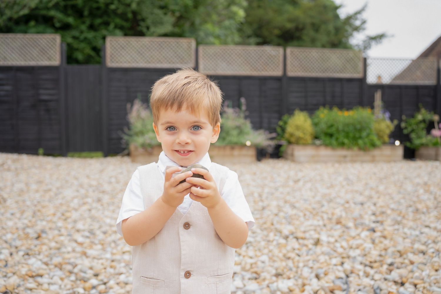 A young boy in a white outfit holds a small object while standing on a gravel path in a garden.