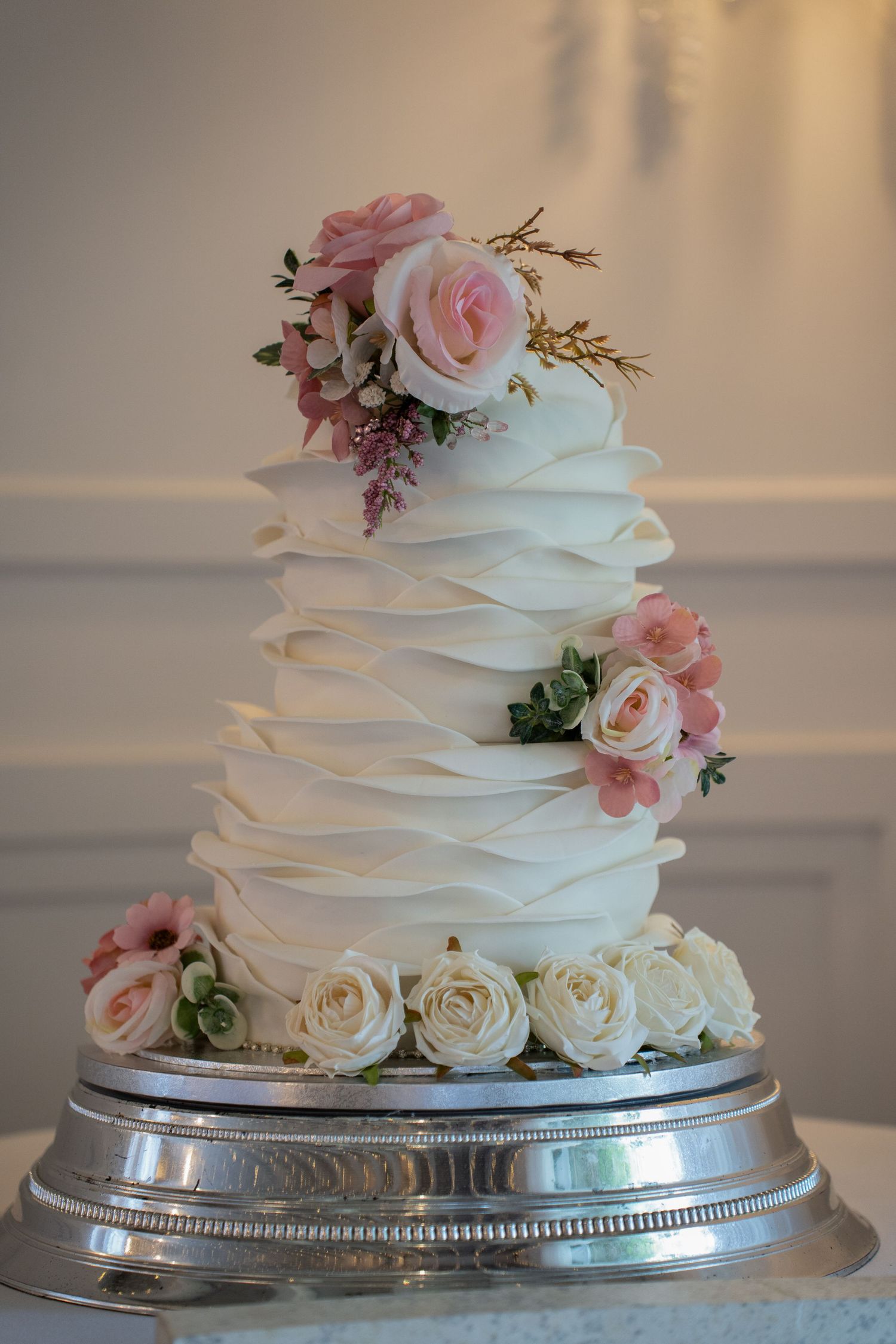 Elegant three-tier white wedding cake with pink and cream roses on silver stand.