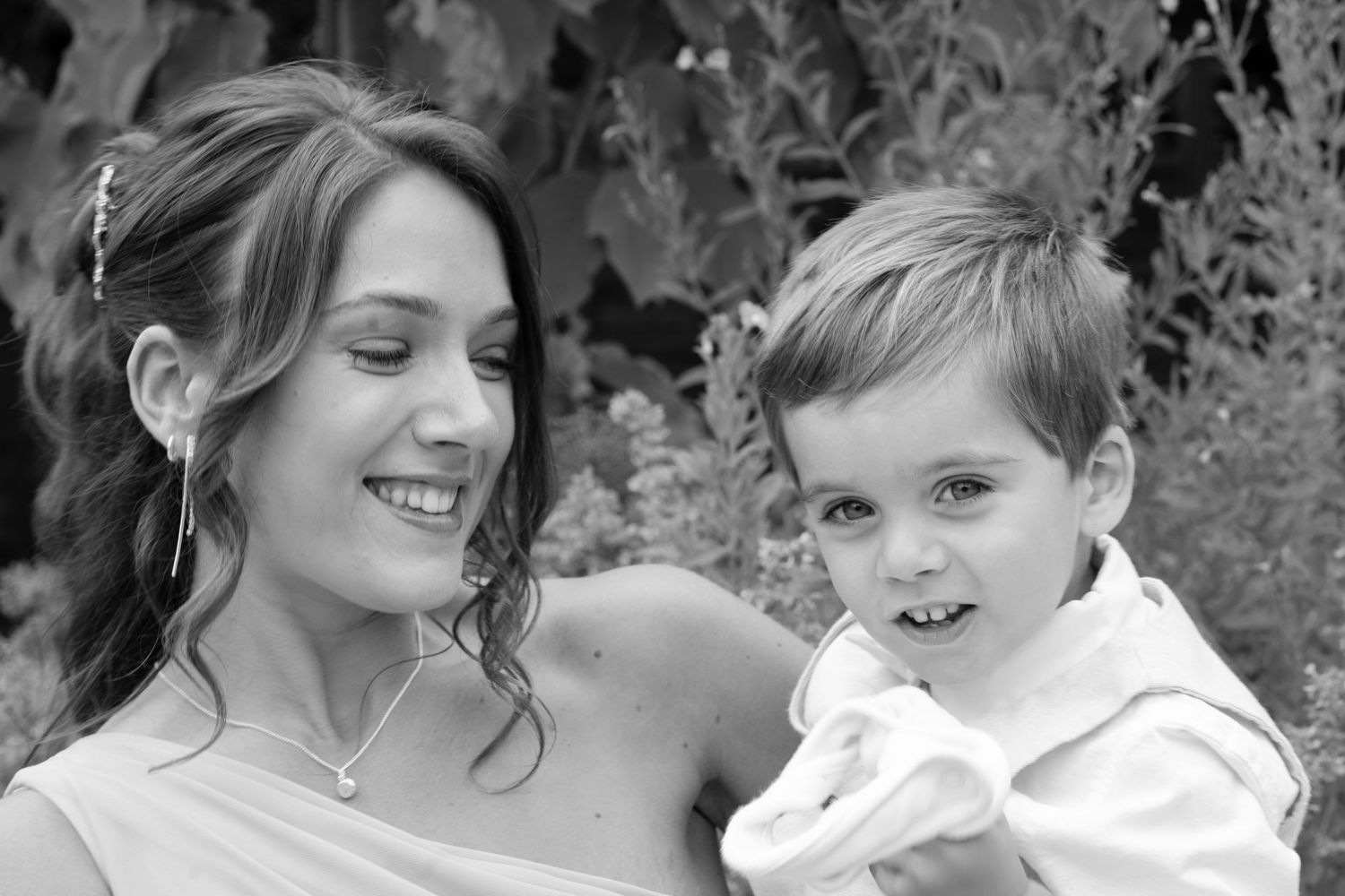 A smiling woman holds a young boy in a white outfit in a black and white portrait photo.