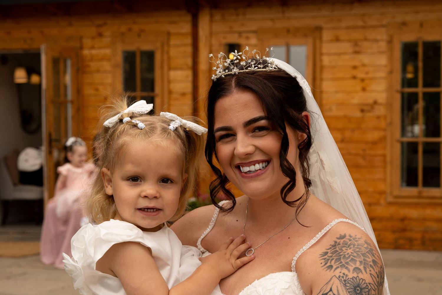 Smiling bride wearing gold crown and veil holding young blonde flower girl in white dress.