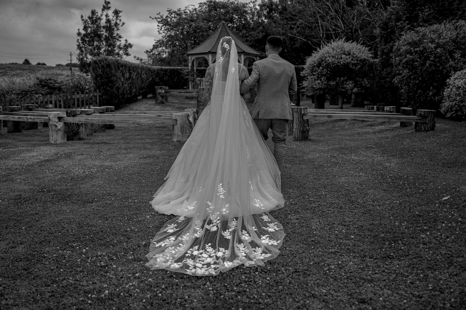 Black and white photo of a bride in a lace-trimmed cathedral veil walking with groom in garden.