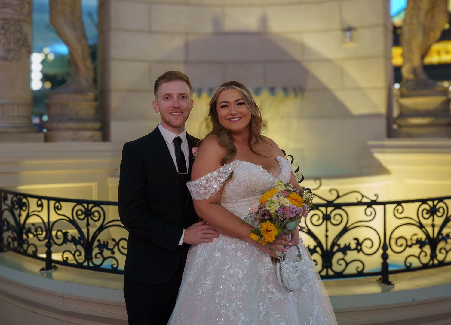 Smiling newlyweds pose together on ornate balcony, bride holding yellow sunflower wedding bouquet.