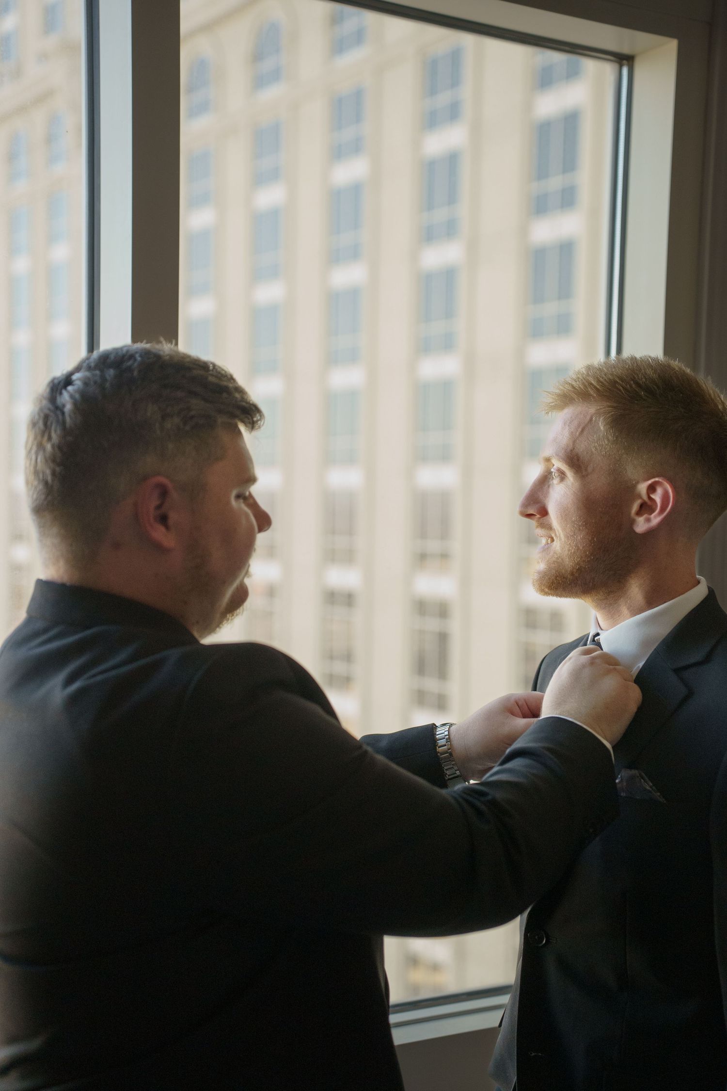 Two men in dark suits adjusting a tie near a large window with city building views.