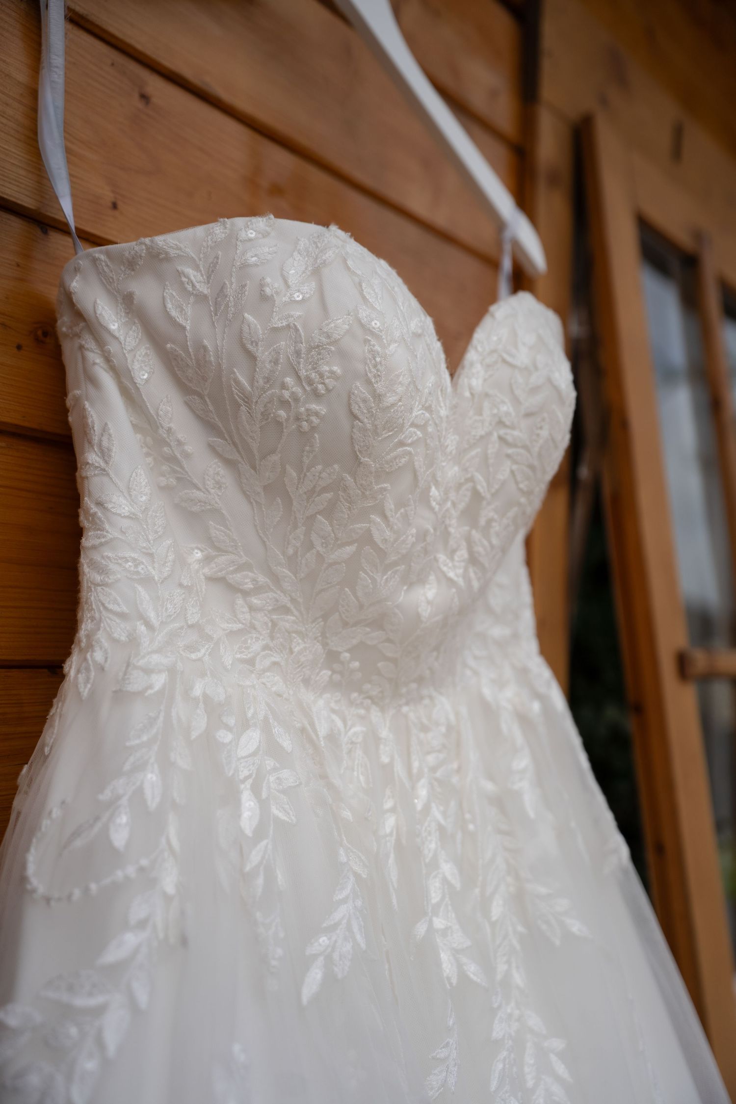 Elegant white lace A-line wedding dress with floral embroidery hanging on wooden wall.