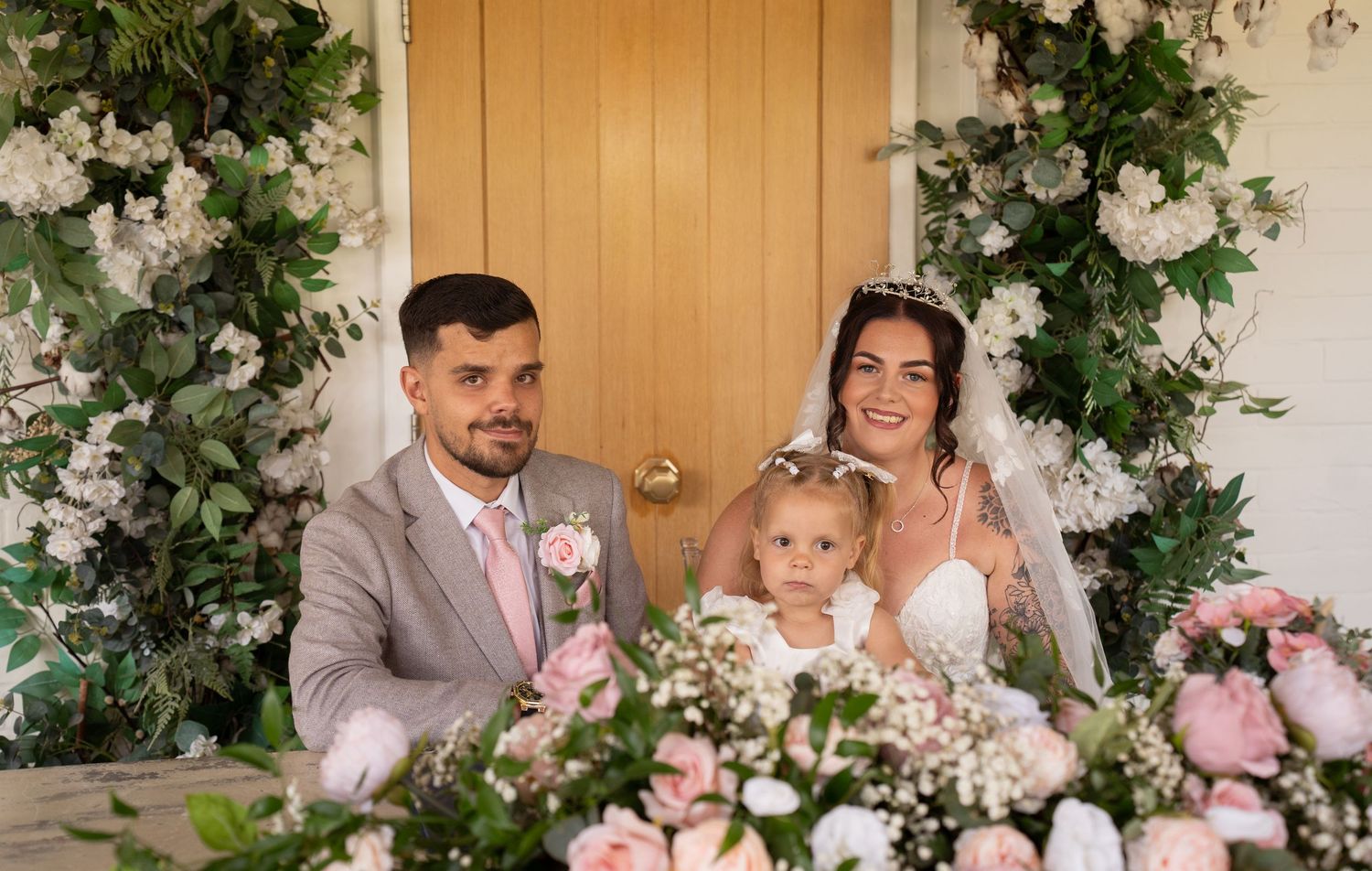 Happy couple with young child seated at wedding ceremony table surrounded by pink floral arrangements.