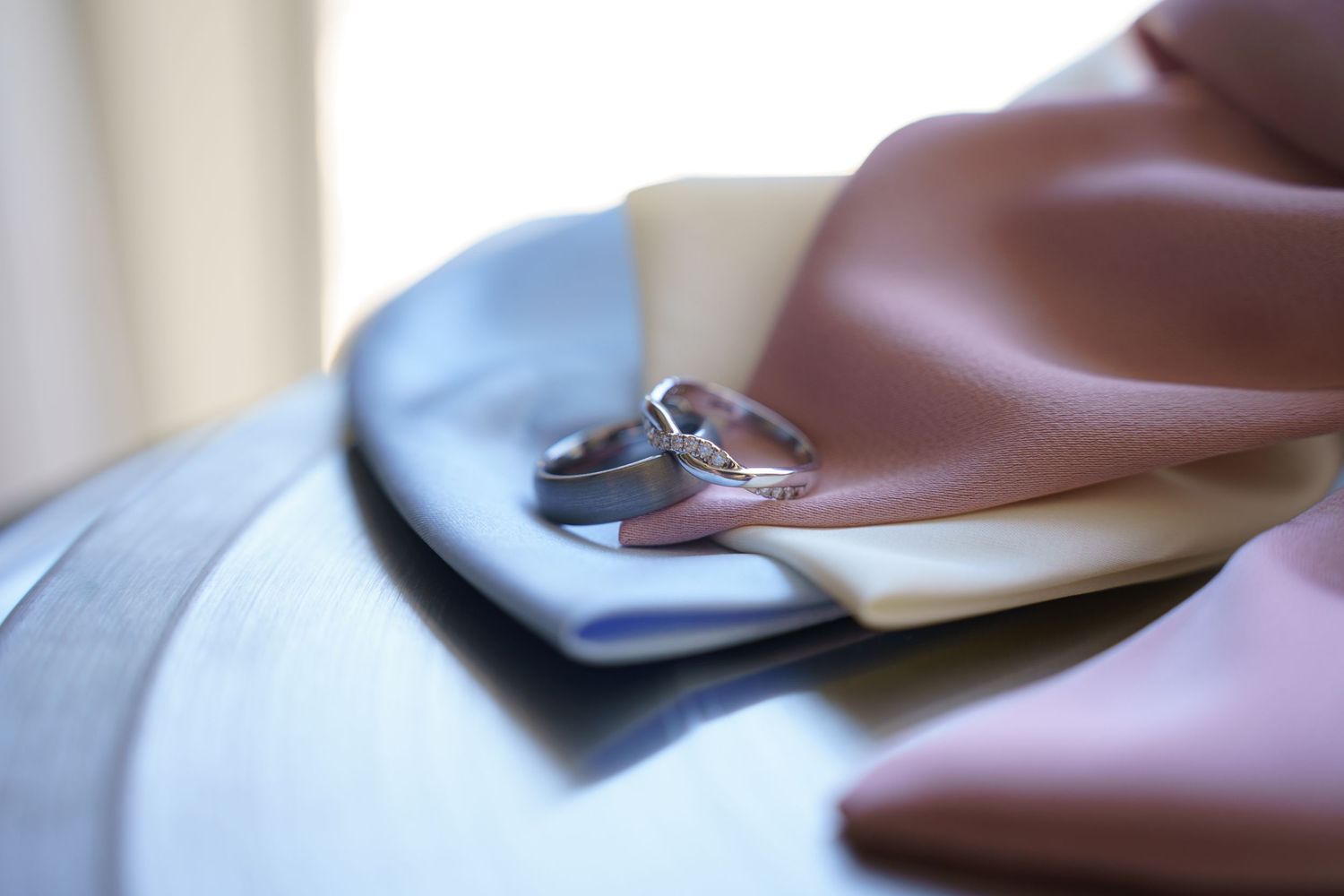 Two silver wedding bands resting on colorful fabric swatches in blue, cream, and pink tones.
