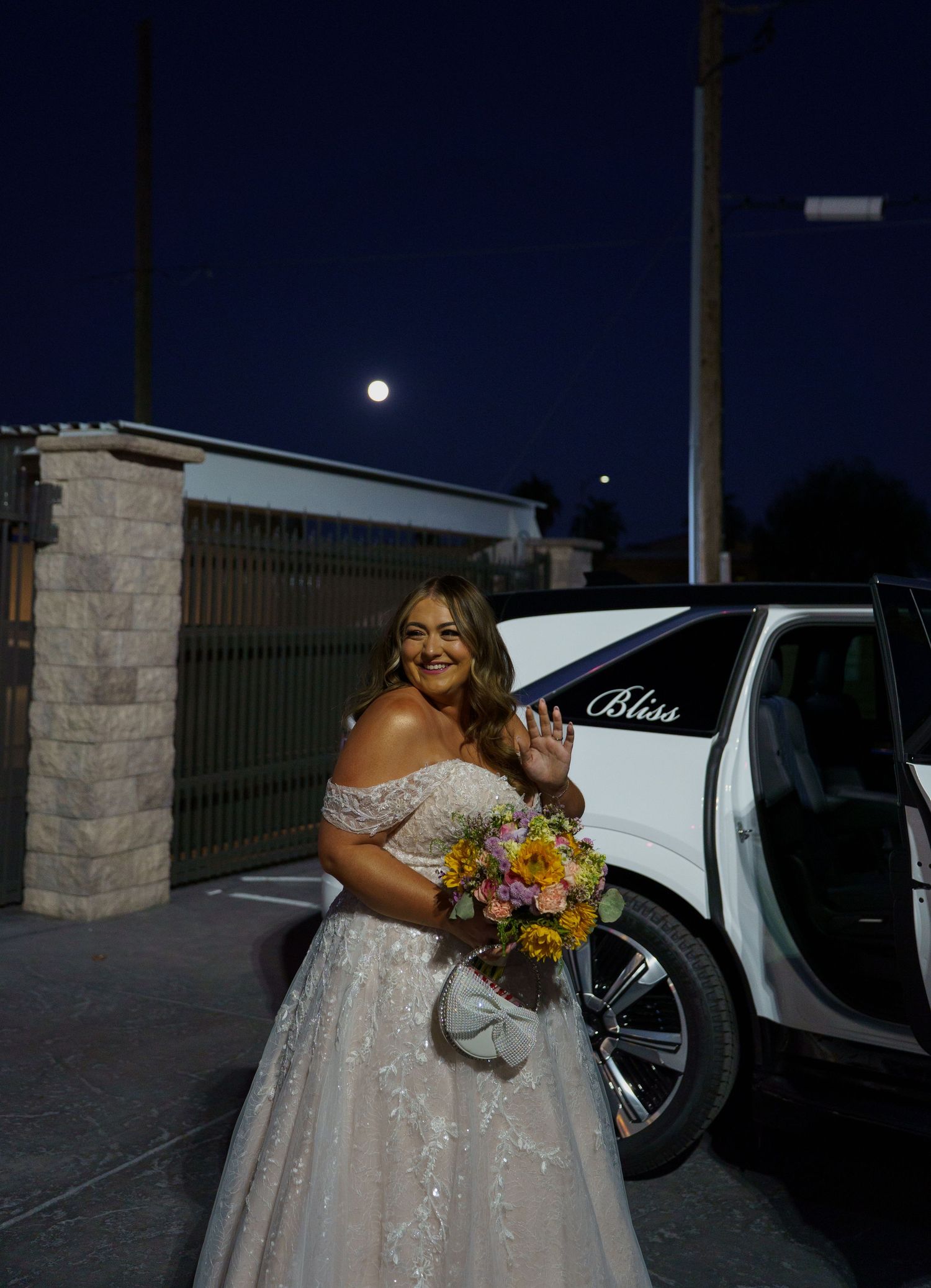 Bride in lace off-shoulder gown waves beside white Bliss limo under a full moon at night.