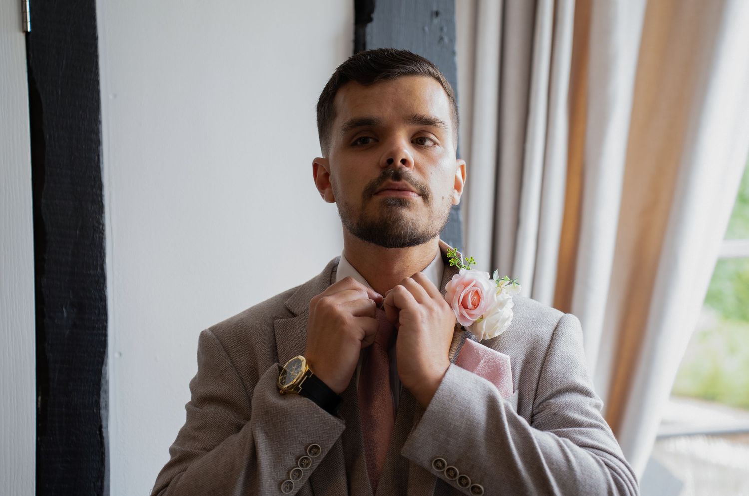 Groom in beige suit adjusting tie with boutonniere near bright window.