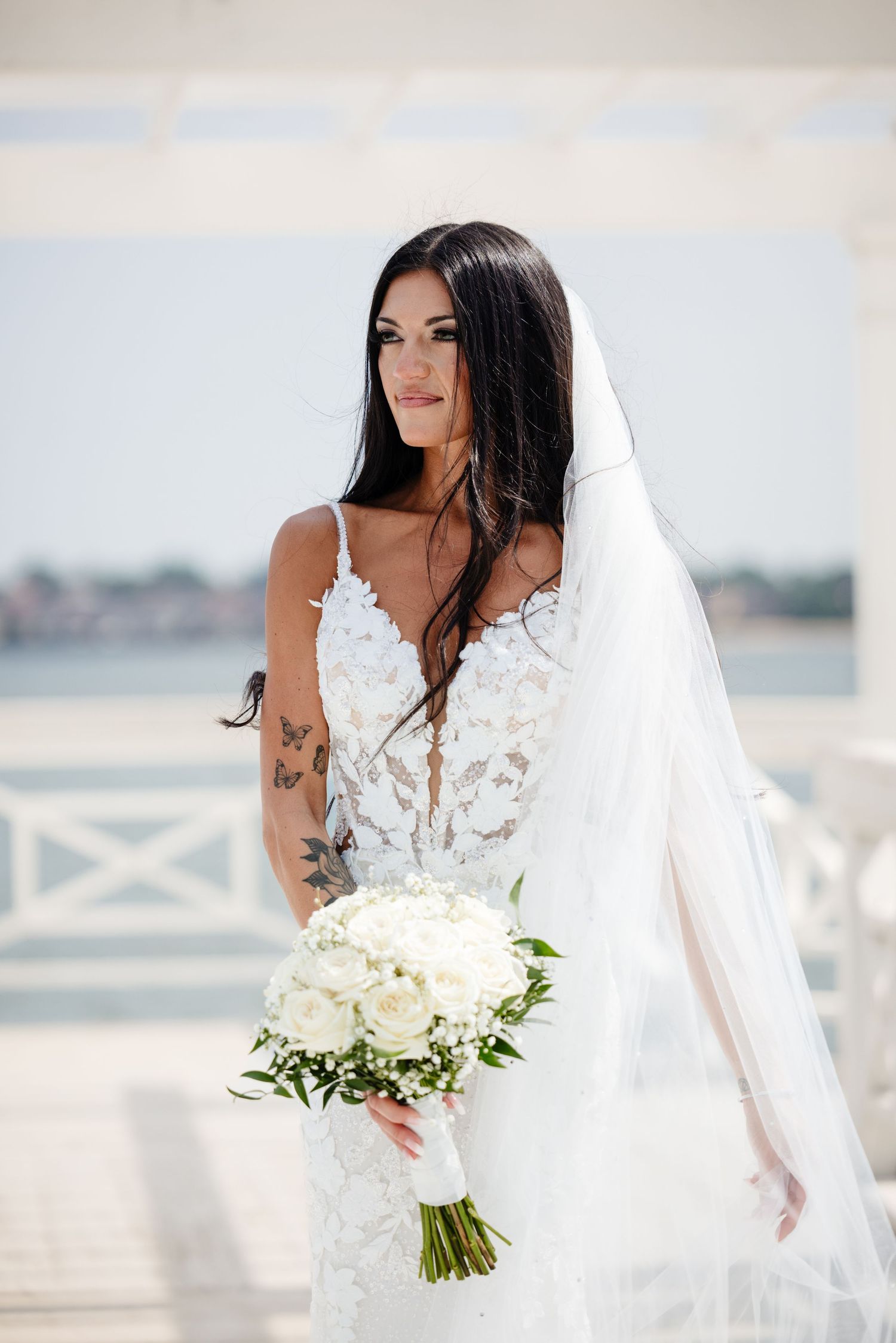 Bride in lace gown holds white bouquet while posing on waterfront terrace.