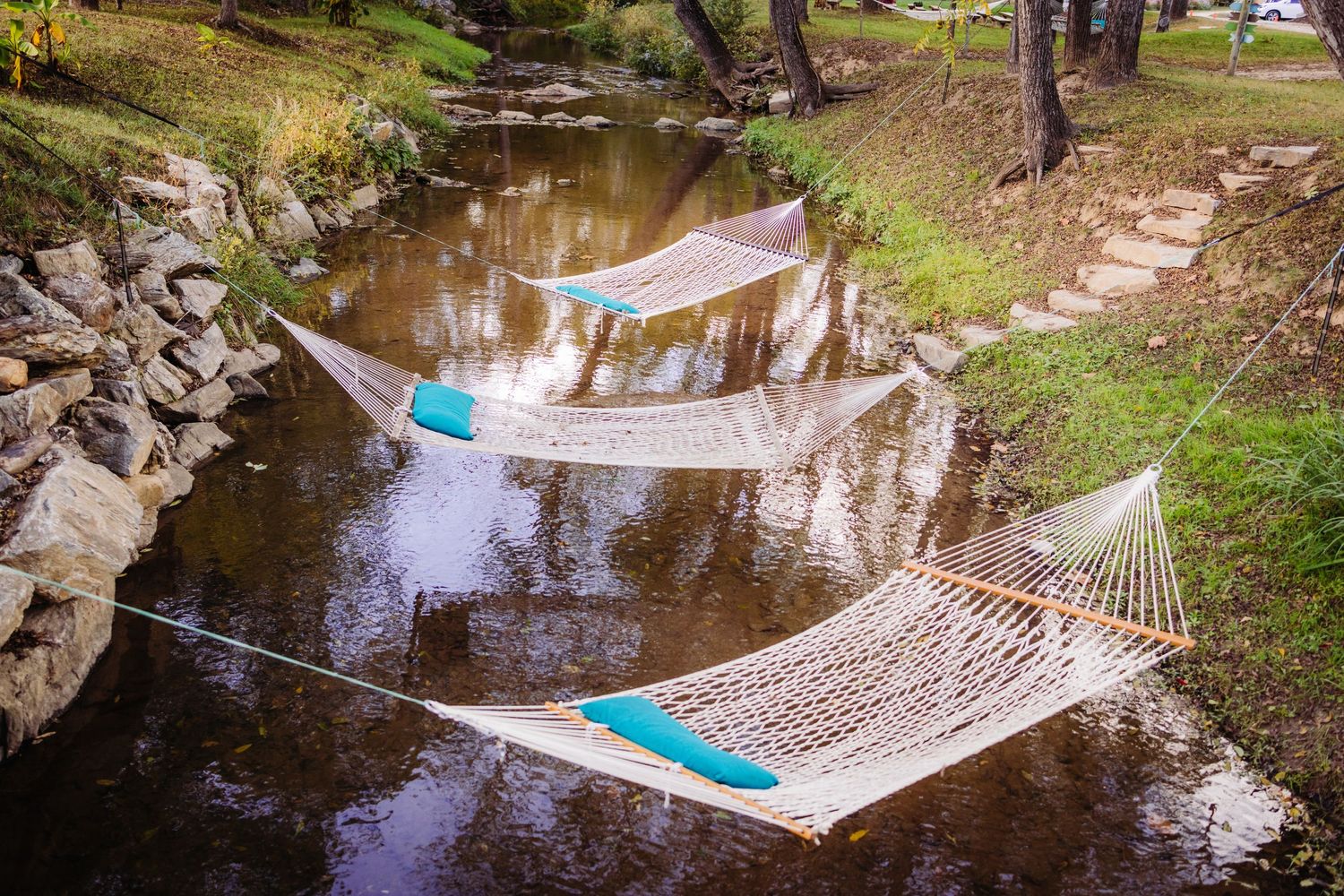 A hammock with turquoise pillows suspended over a reflective stream in a wooded setting.