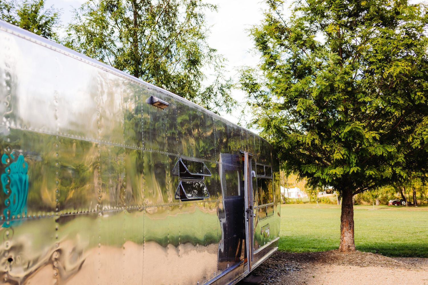 A shiny vintage Airstream trailer parked in a sunny outdoor setting with green trees in the background.