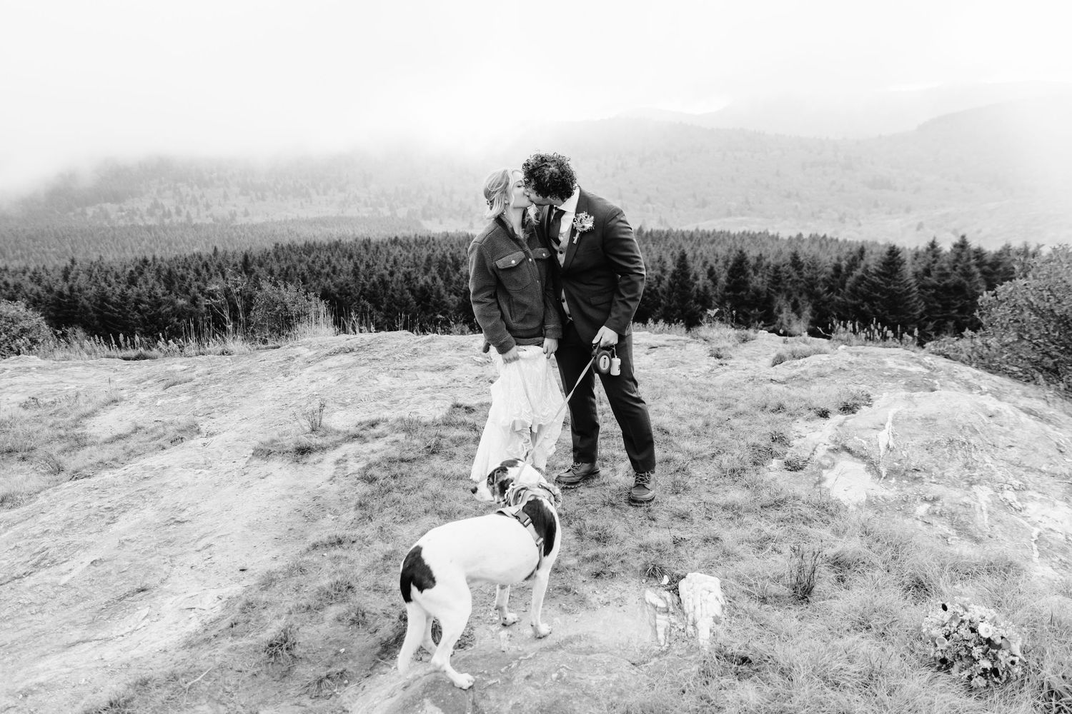 Couple embraces on mountain summit with two dogs against scenic forest landscape in black and white.