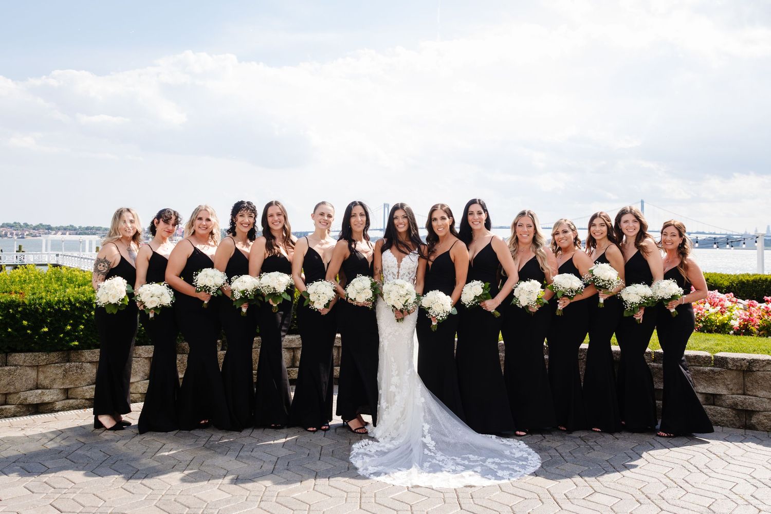 Wedding party of bridesmaids in black dresses poses with bride in white gown by waterfront.