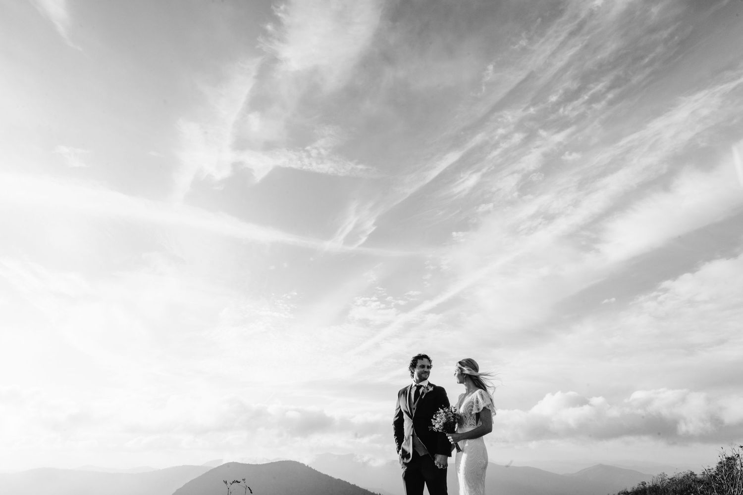 Couple embraces on a scenic mountain ridge during sunset with dramatic cloudy skies in the background.