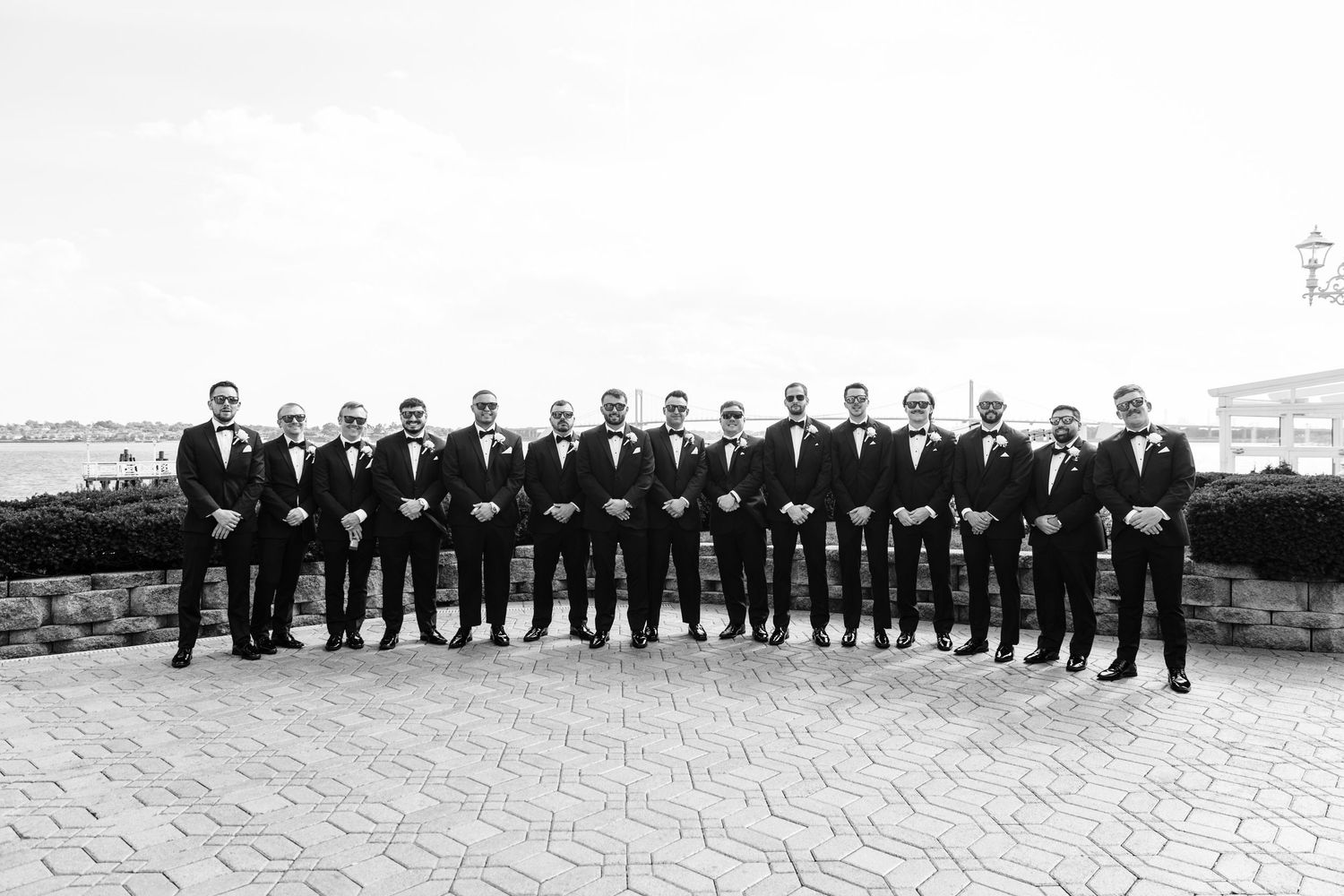 Large wedding party of groomsmen dressed in black suits stand together outdoors.