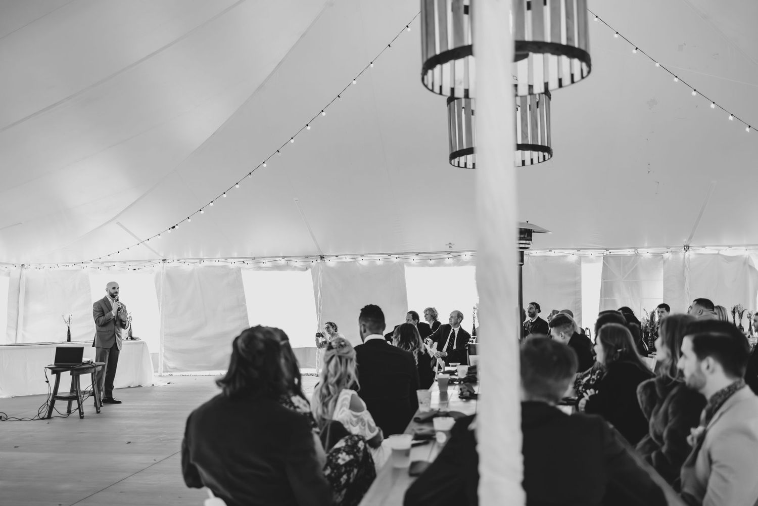 Wedding guests gather under a white tent with hanging lights and chandeliers.
