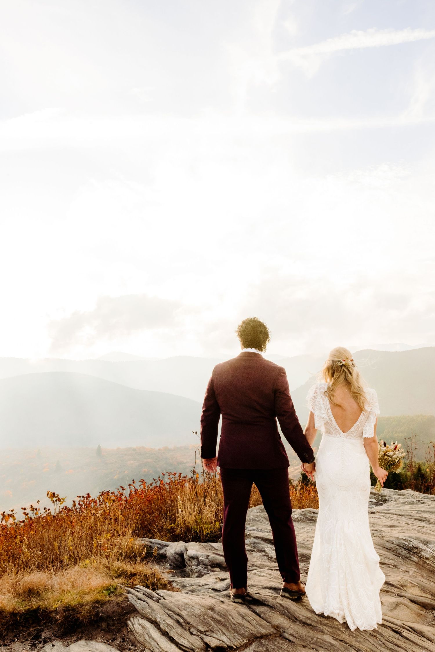 Couple stands hand in hand overlooking mountain vista at sunset.