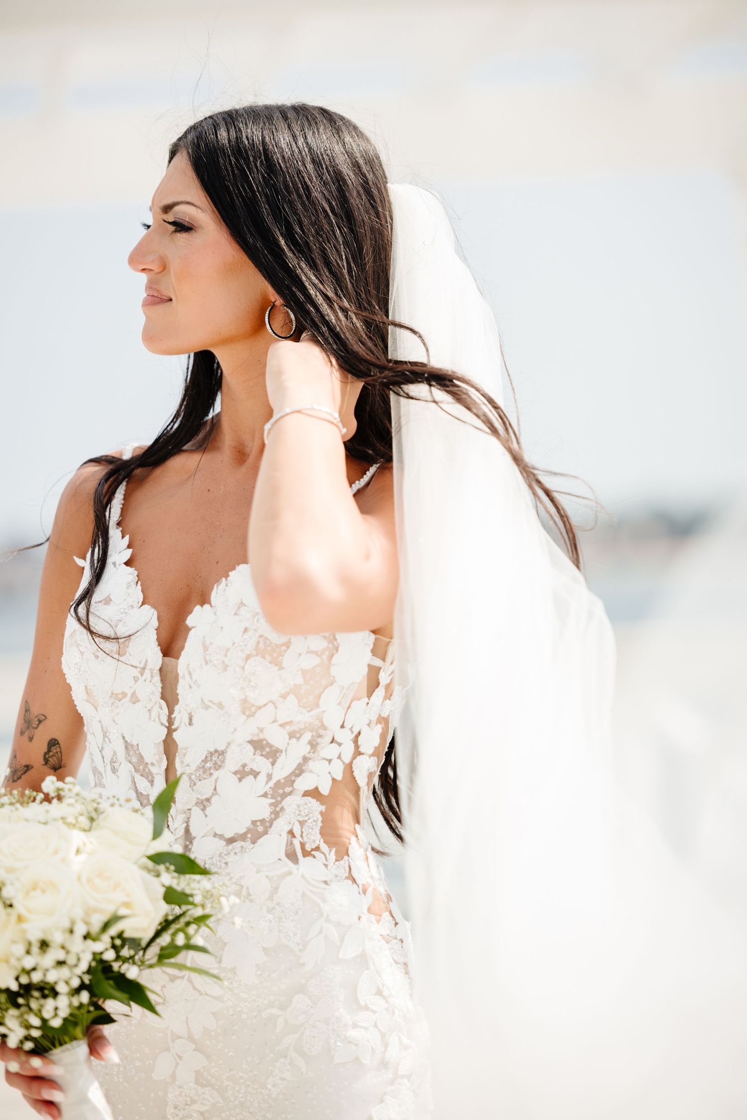Side profile of bride in lace dress holding bouquet with flowing veil in sunlight.