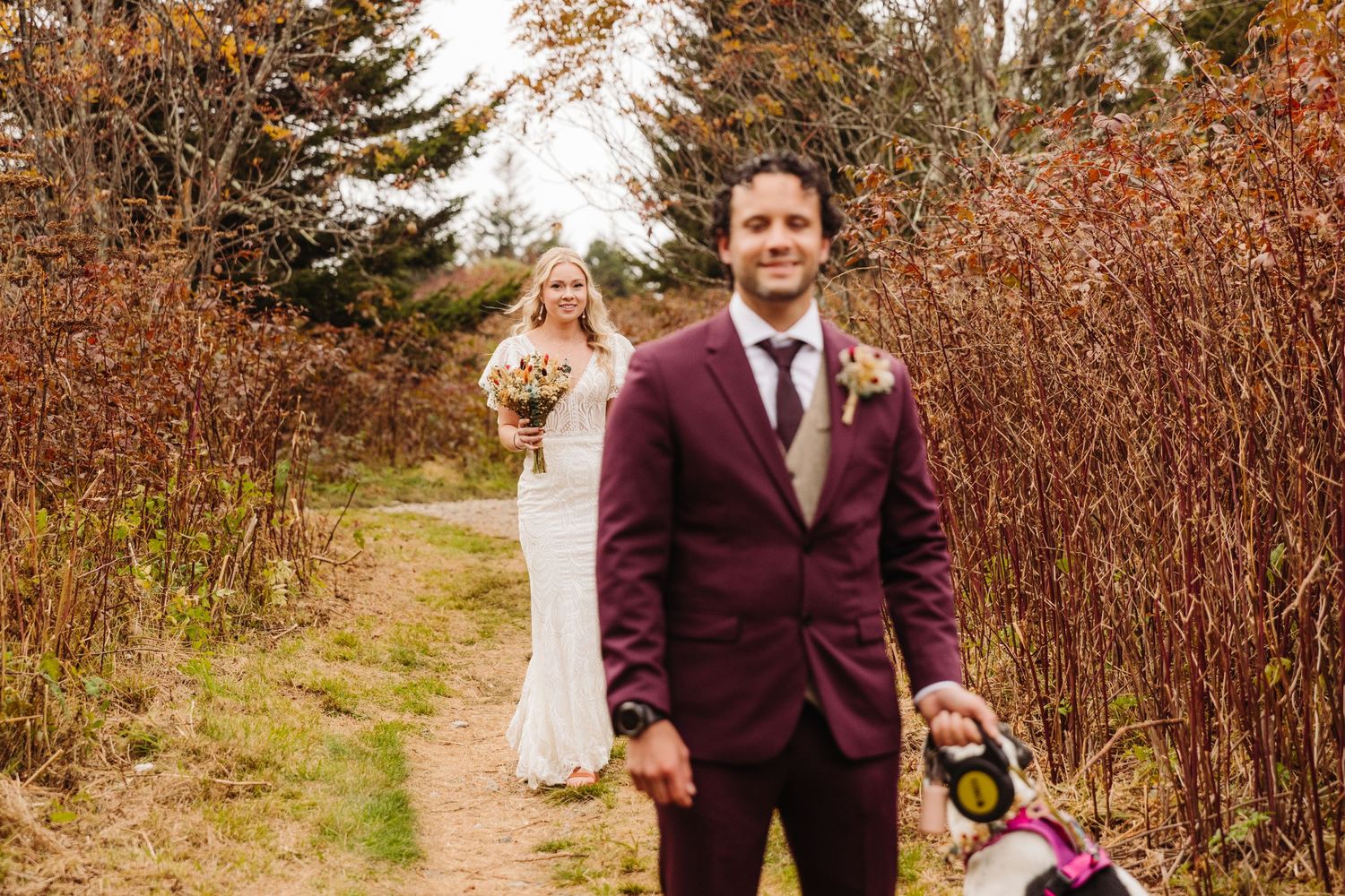 Fall wedding photo with burgundy suit and white dress on nature path.