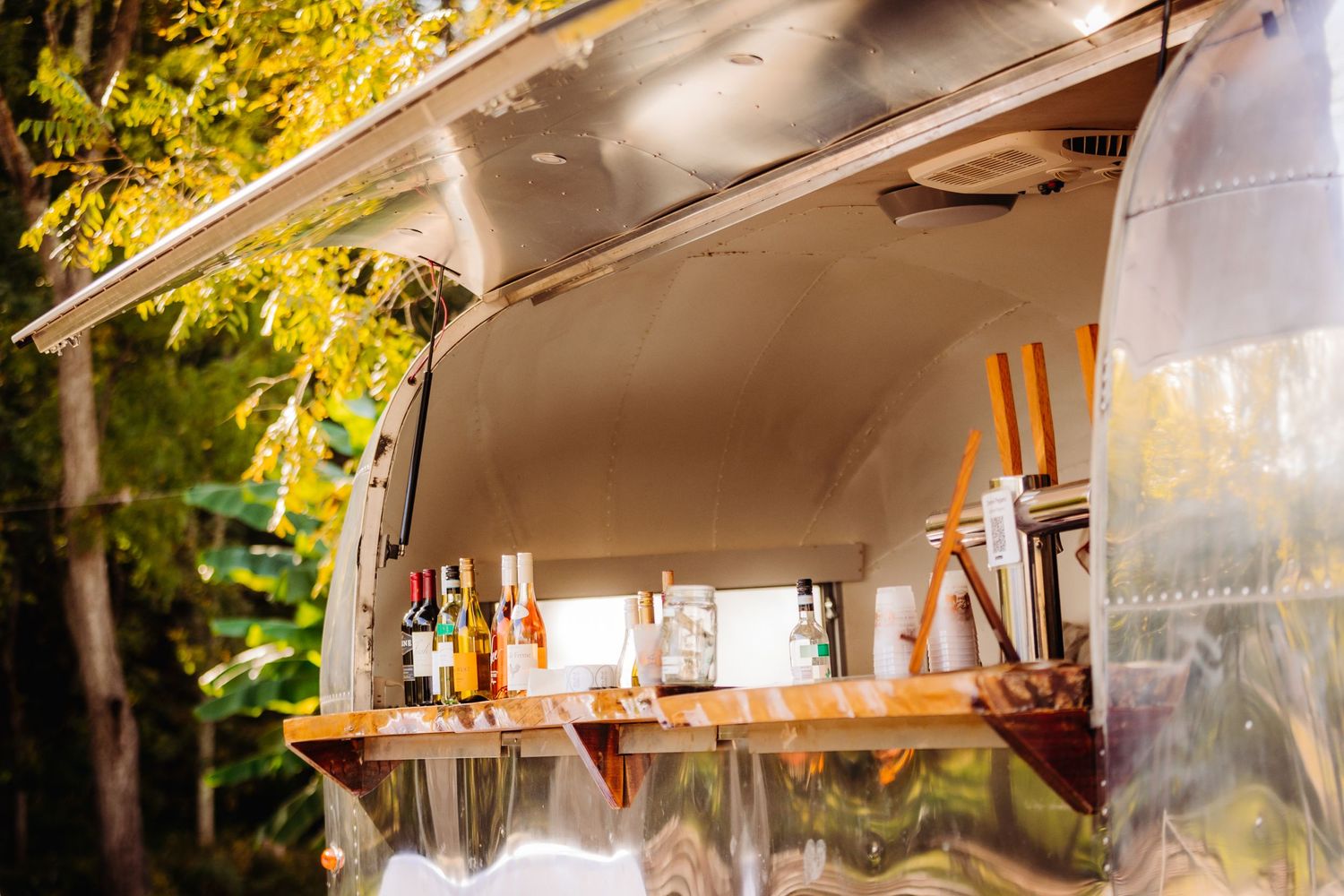 A vintage Airstream trailer's bar window displaying glass bottles and cocktail supplies.