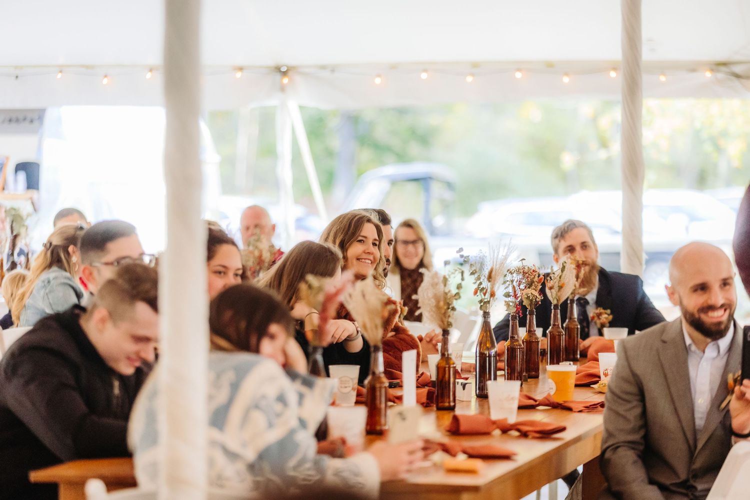 Wedding reception guests enjoy drinks and conversation at a long table under a tent.