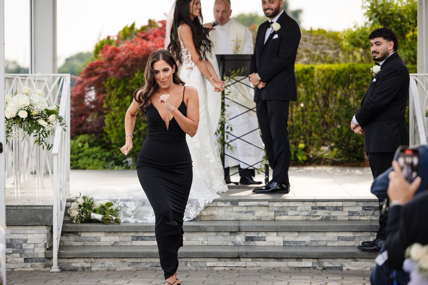 Outdoor wedding ceremony setup with white columns and flowering bushes in the background.