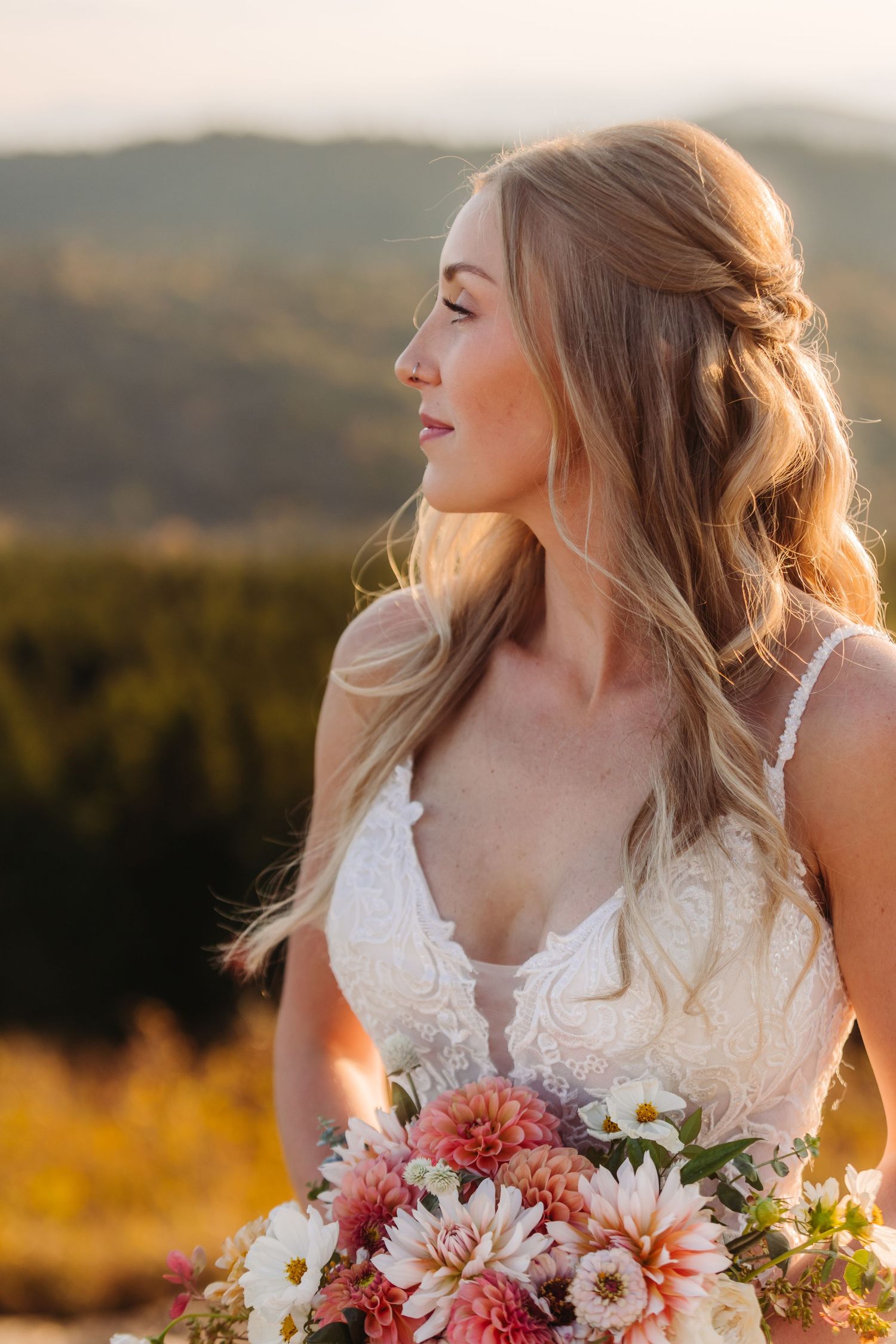 Bride in lace dress holds bouquet while gazing at mountain sunset view.