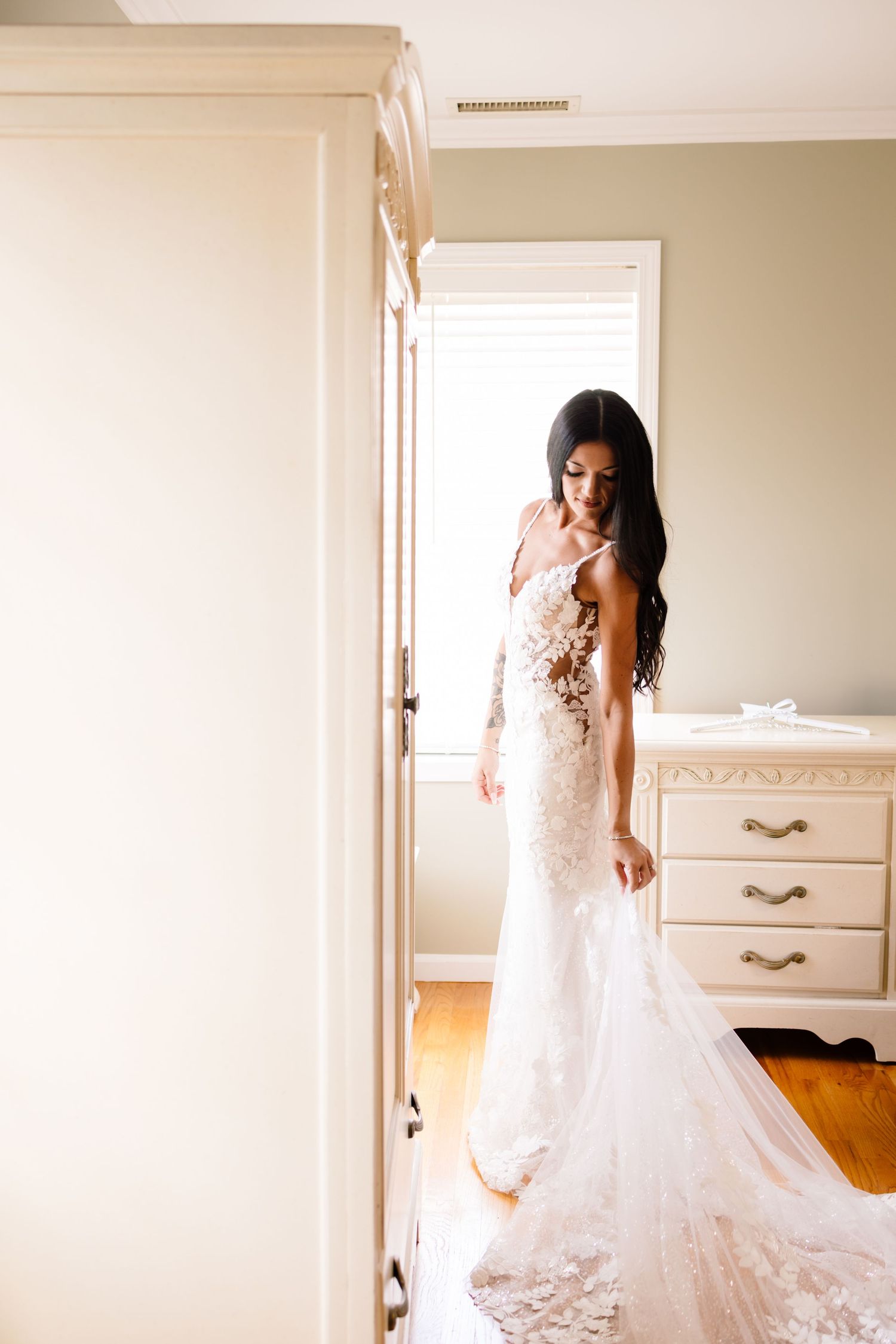 Elegant bride in fitted lace wedding gown with long train standing in sunlit room.