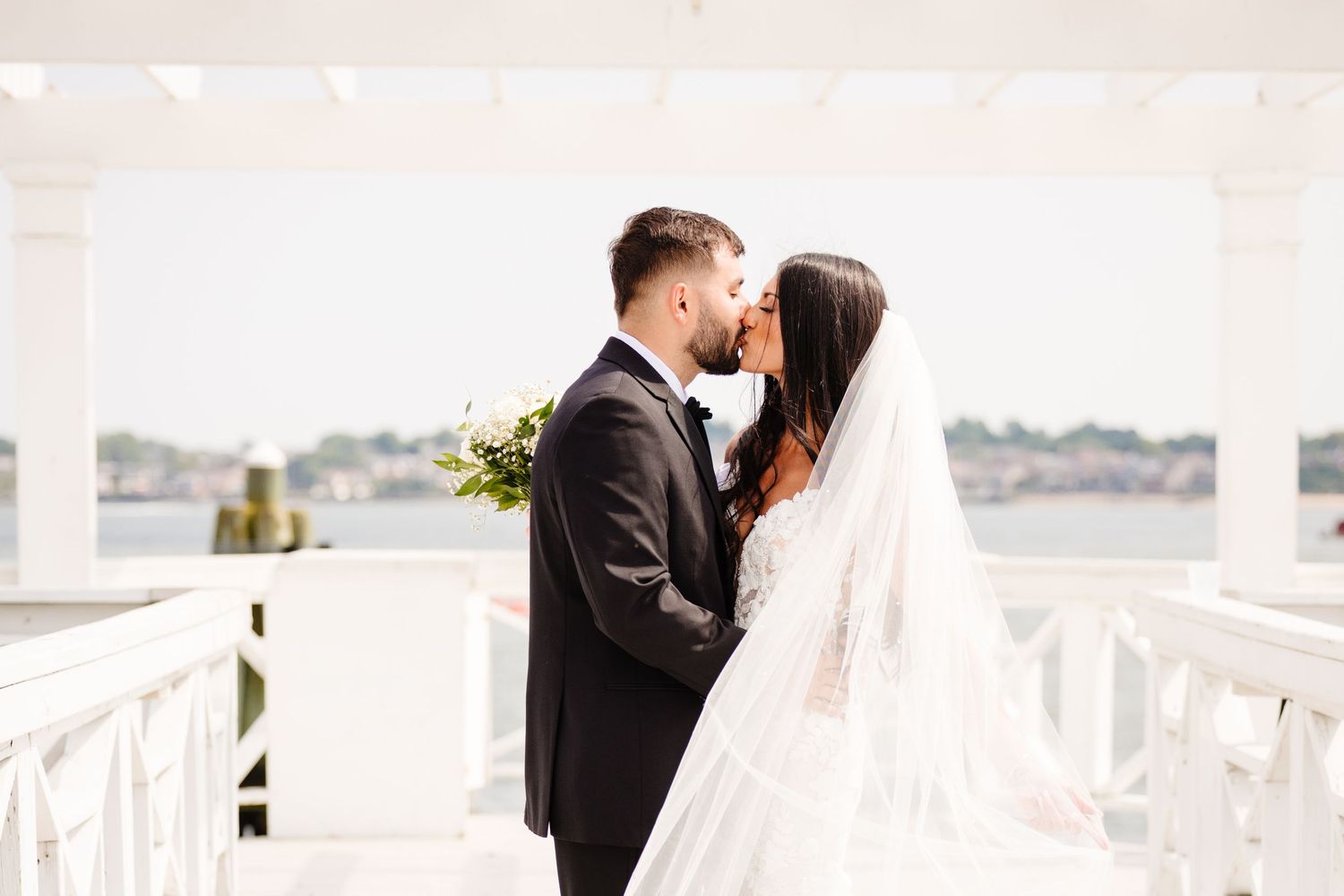 Couple shares a tender moment on their wedding day under a white pergola by the water.