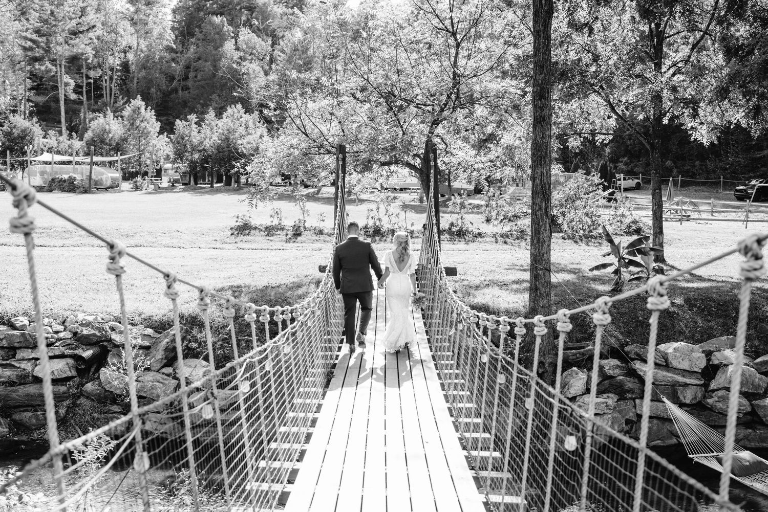 A black and white photo of a couple crossing a rustic rope bridge in nature.