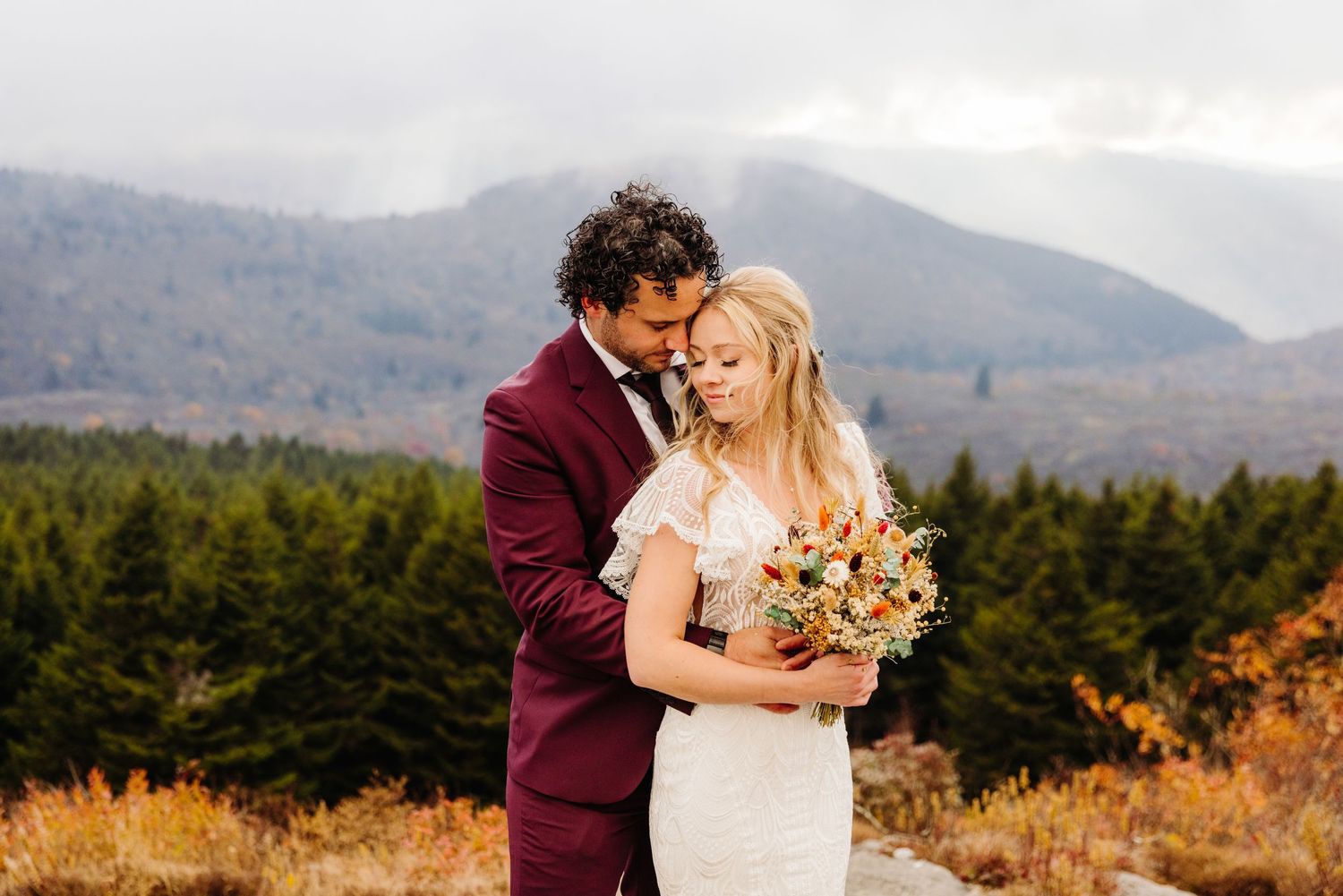 Romantic autumn wedding portrait with burgundy suit against mountain landscape.