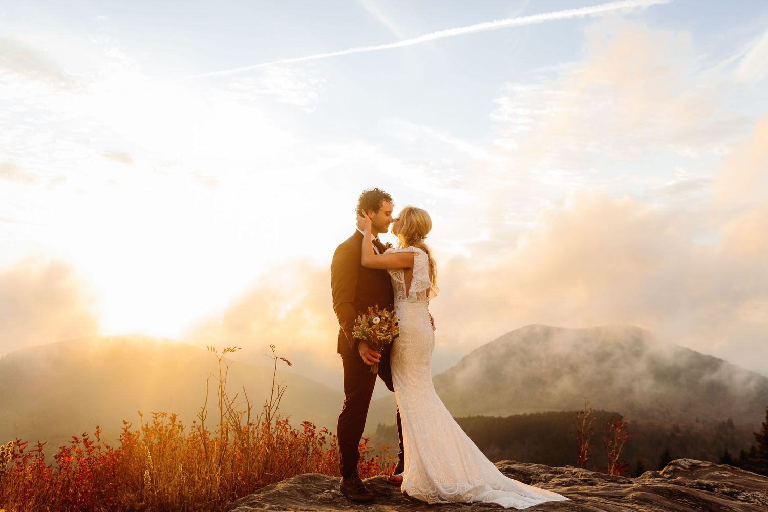 A couple embraces on a mountaintop during golden hour with misty peaks in the distance.